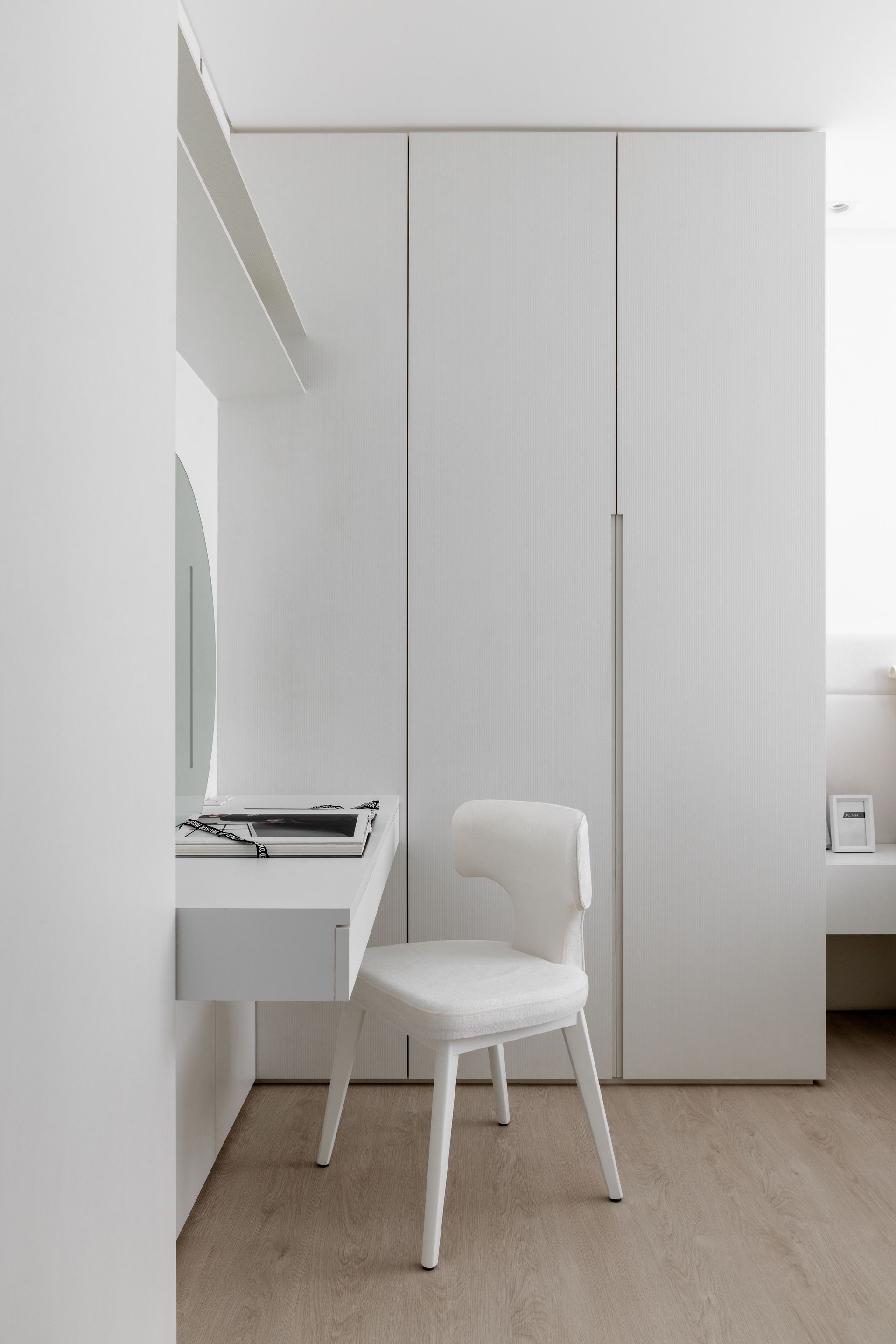 Toucador minimalista branco com uma cadeira ao lado de um grande guarda-roupa em um quarto de cores claras.