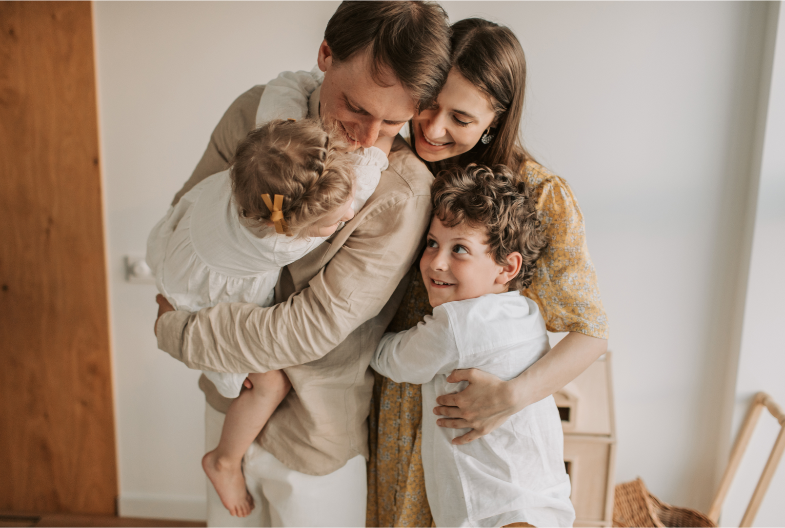 A family hugging indoors, with two adults embracing two smiling children in a cozy room.