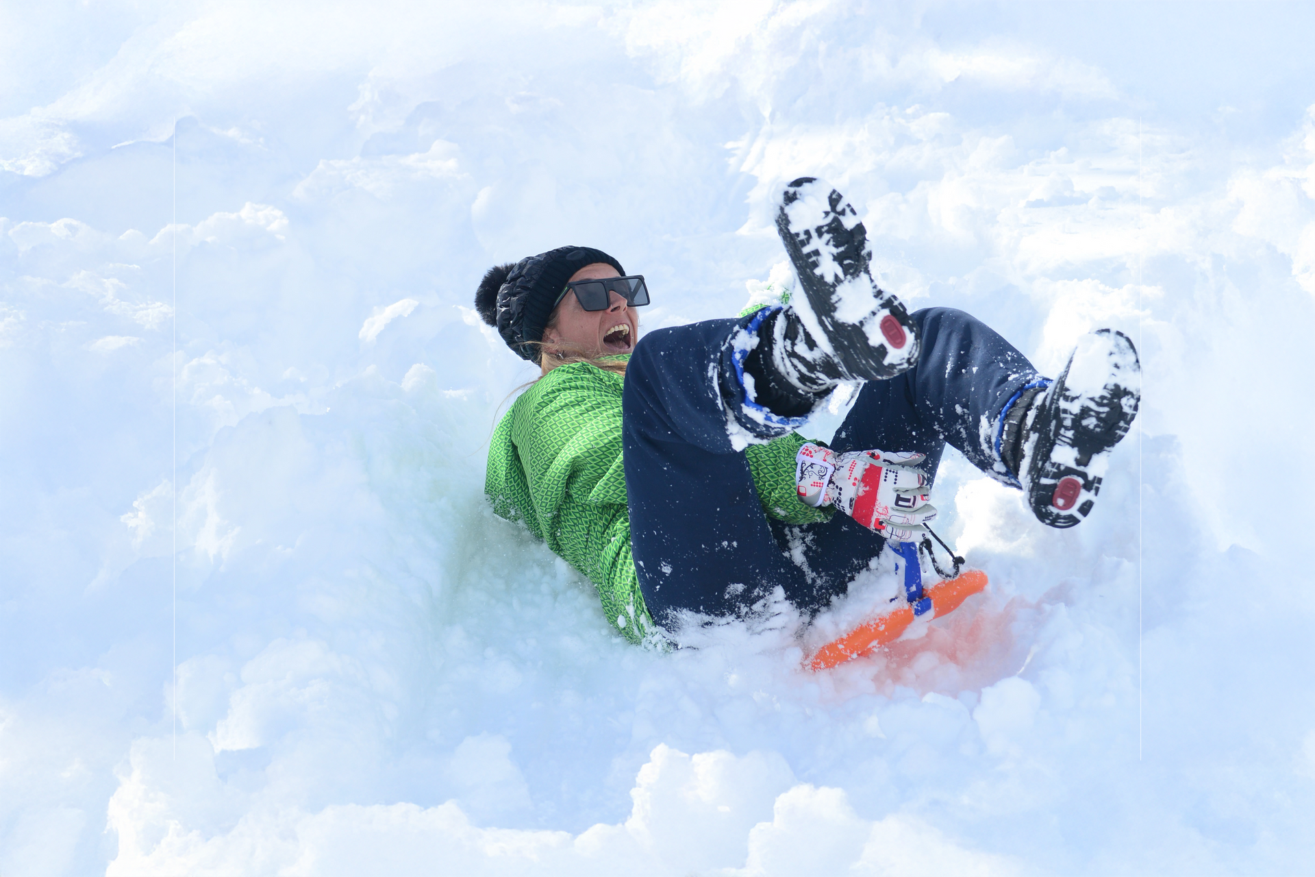 Persona con gafas de sol y gorro dando volteretas en la nieve fresca, cerca de un trineo de plástico.
