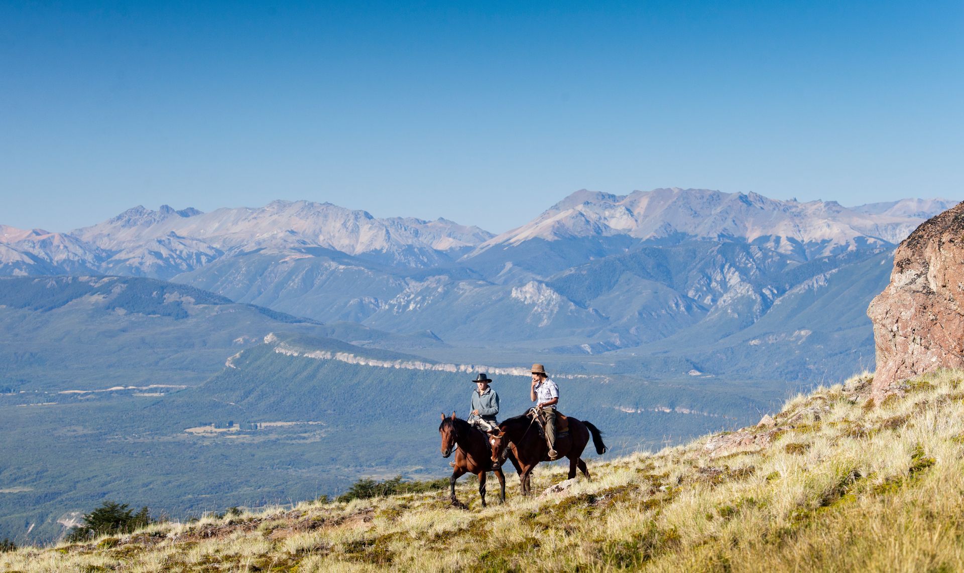 Dos personas a caballo cabalgando sobre una colina cubierta de hierba, con montañas al fondo bajo un cielo azul.