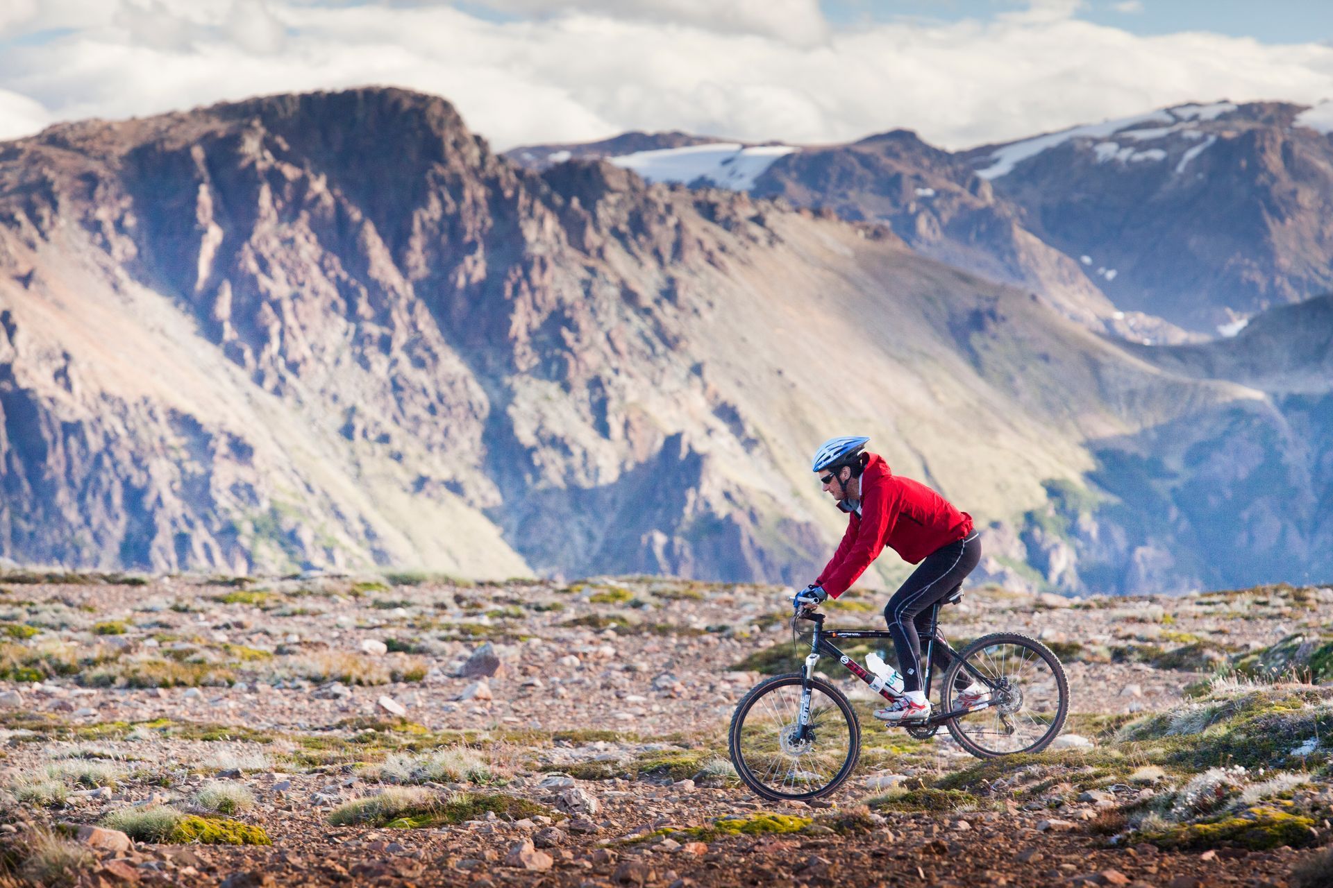 Ciclista de montaña con chaqueta roja circula por terreno rocoso, con montañas al fondo.