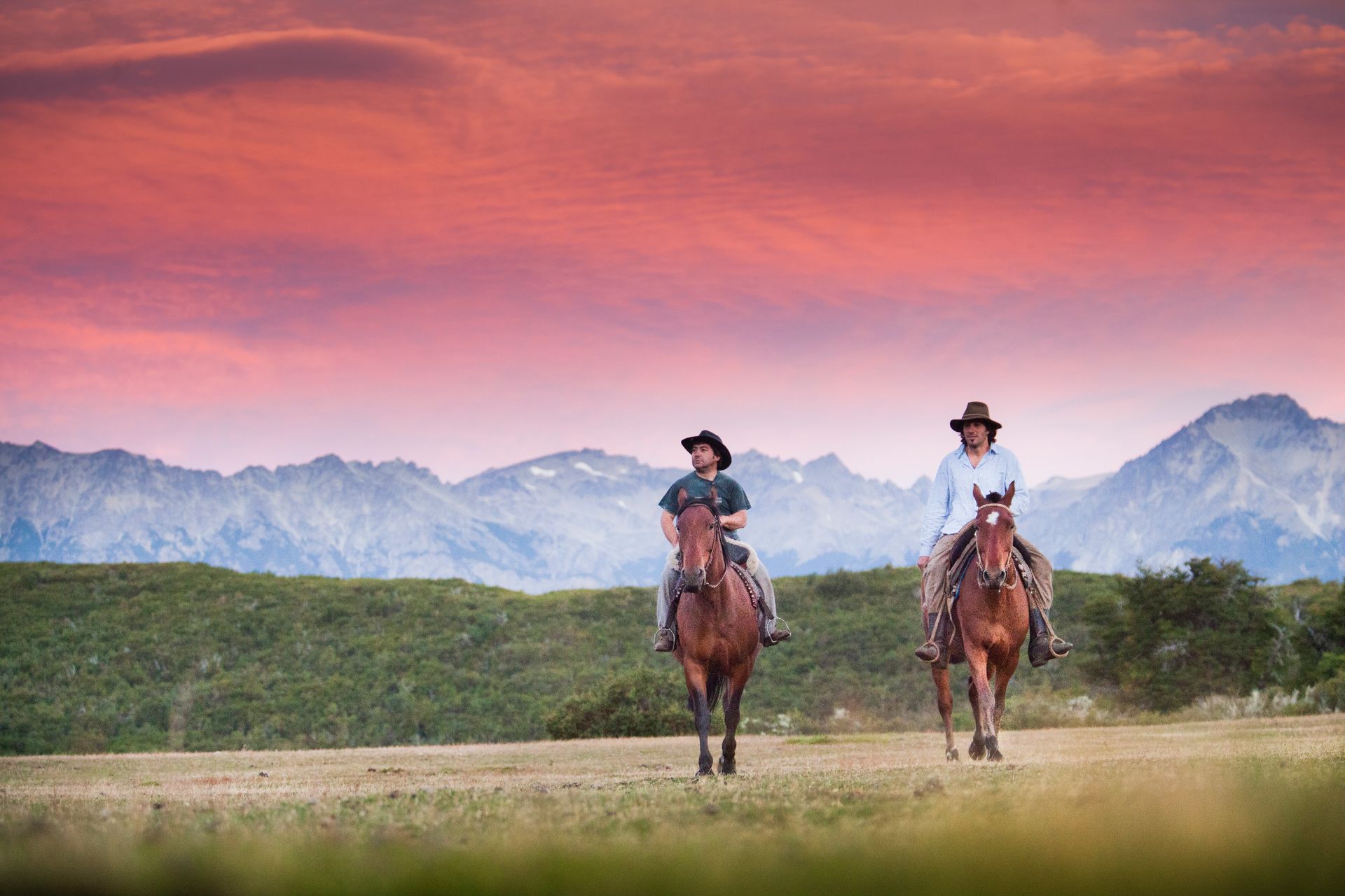 Dos personas a caballo cabalgan por un campo hacia una cadena montañosa bajo un cielo rosa y naranja.