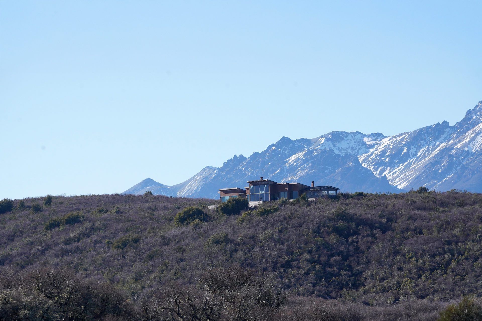 Una casa se encuentra en la cima de una colina, con montañas cubiertas de nieve y un cielo azul en el fondo.