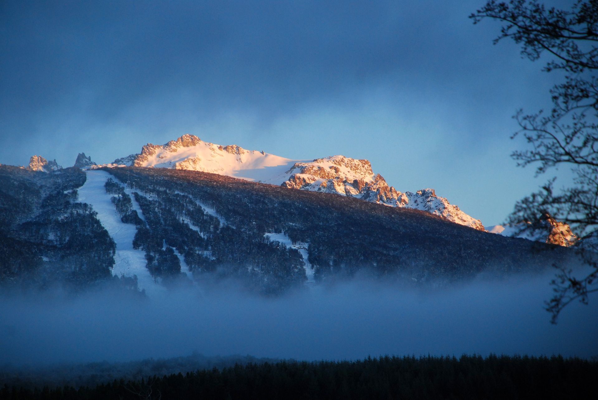 Pico de montaña nevado con árboles y niebla baja bajo un cielo azul al atardecer.