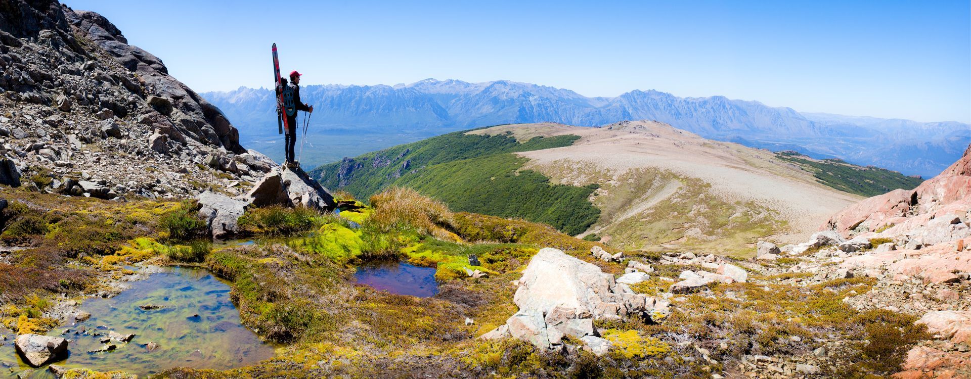 Persona con los brazos levantados en la cima de una montaña, con vistas a un valle y montañas distantes bajo un cielo azul.