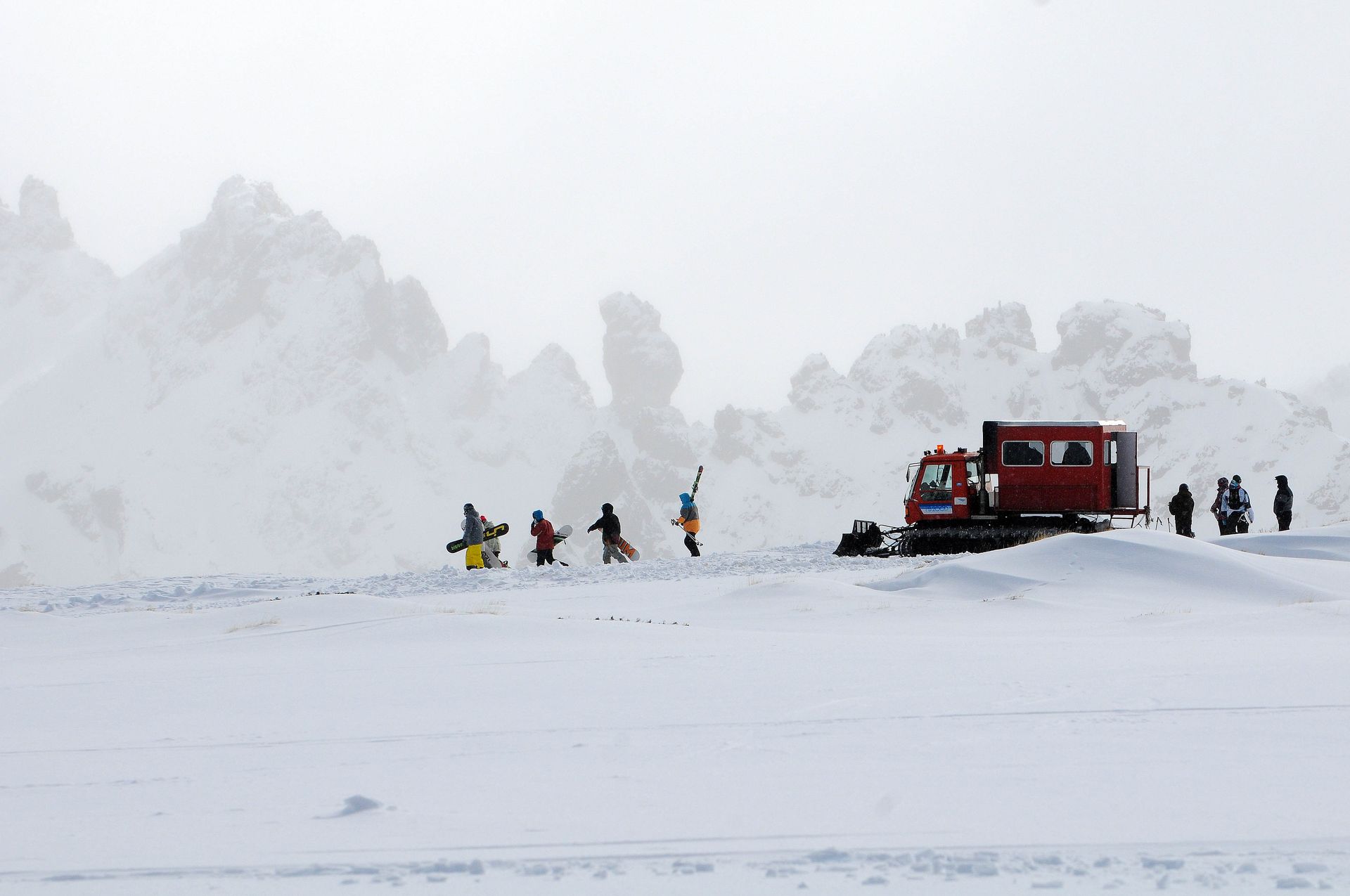 Paisaje nevado con gente y un vehículo para nieve. Personas con ropa colorida caminan cerca de un snowcat rojo.