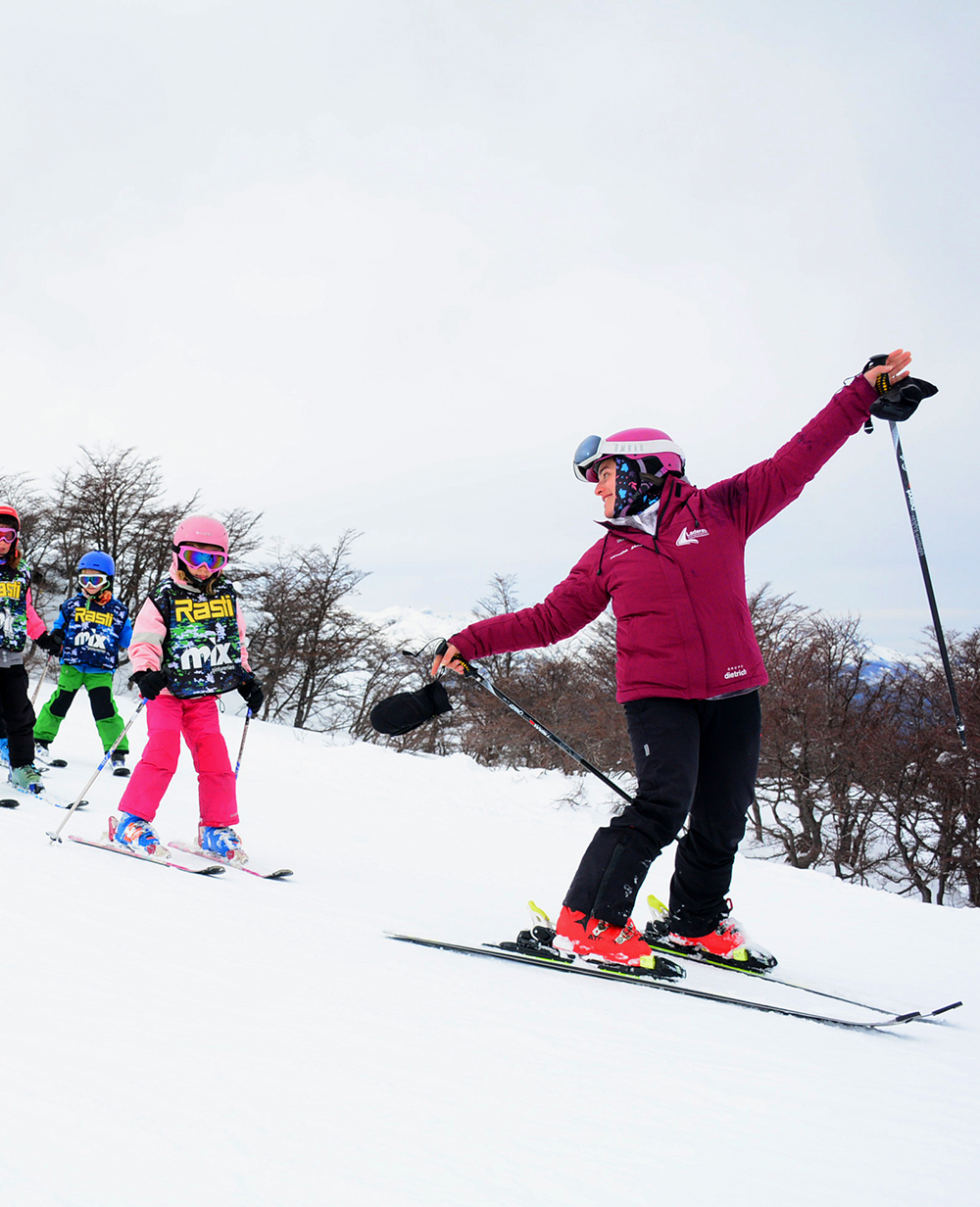 Clase de esquí en grupo: Instructor con chaqueta roja guía a los niños en una pendiente cubierta de nieve; cielo nublado.