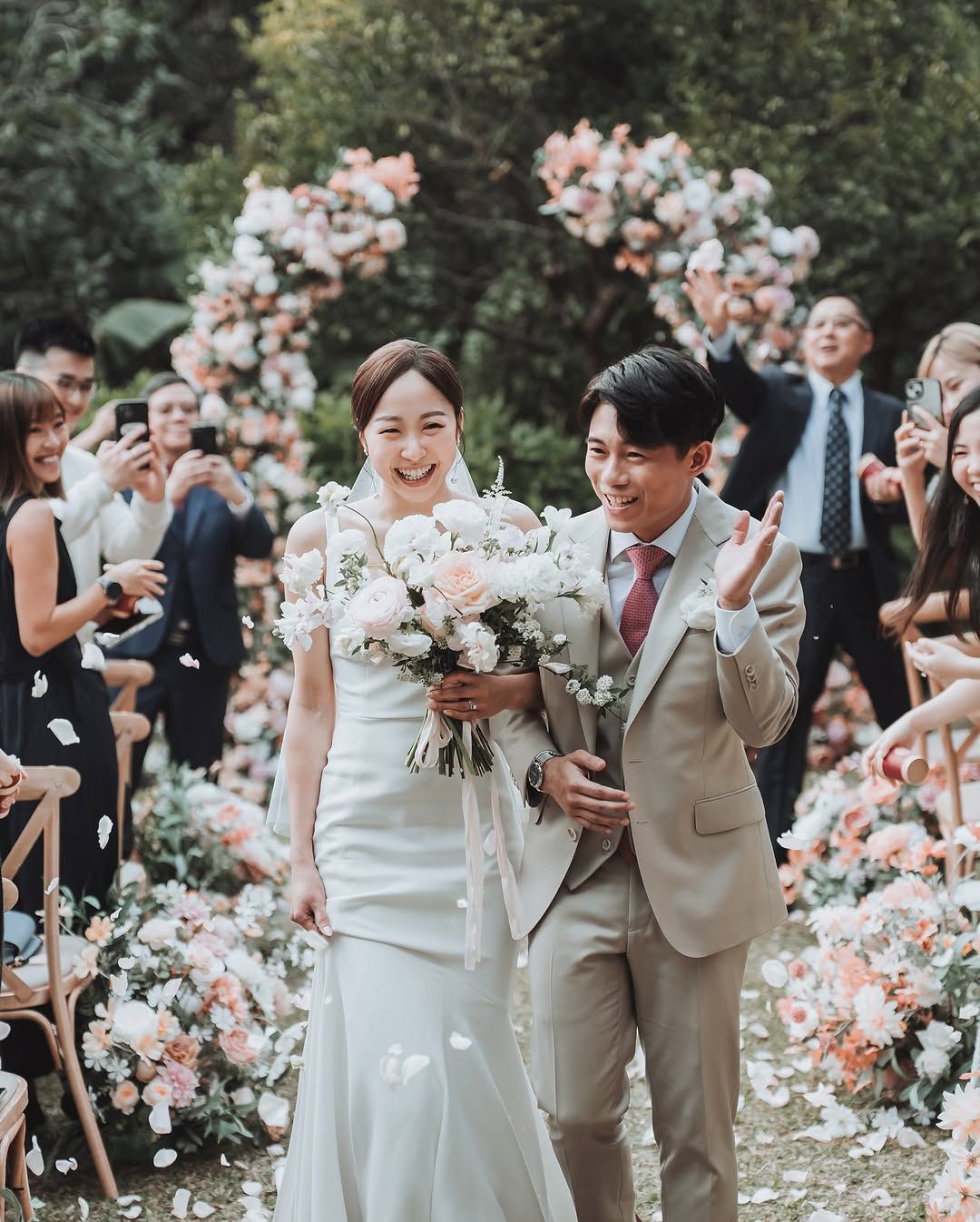 Newlyweds walk down aisle; flowers, guests throwing petals, celebratory smiles.