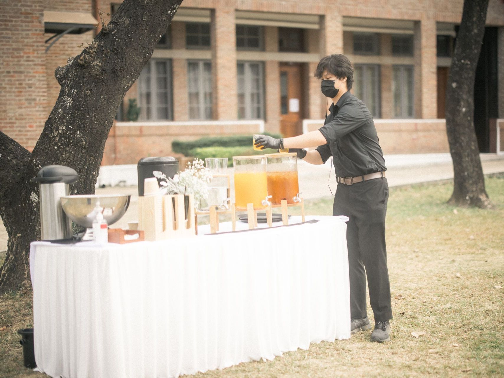 A person in black attire serves drinks at an outdoor buffet, set against a brick building.