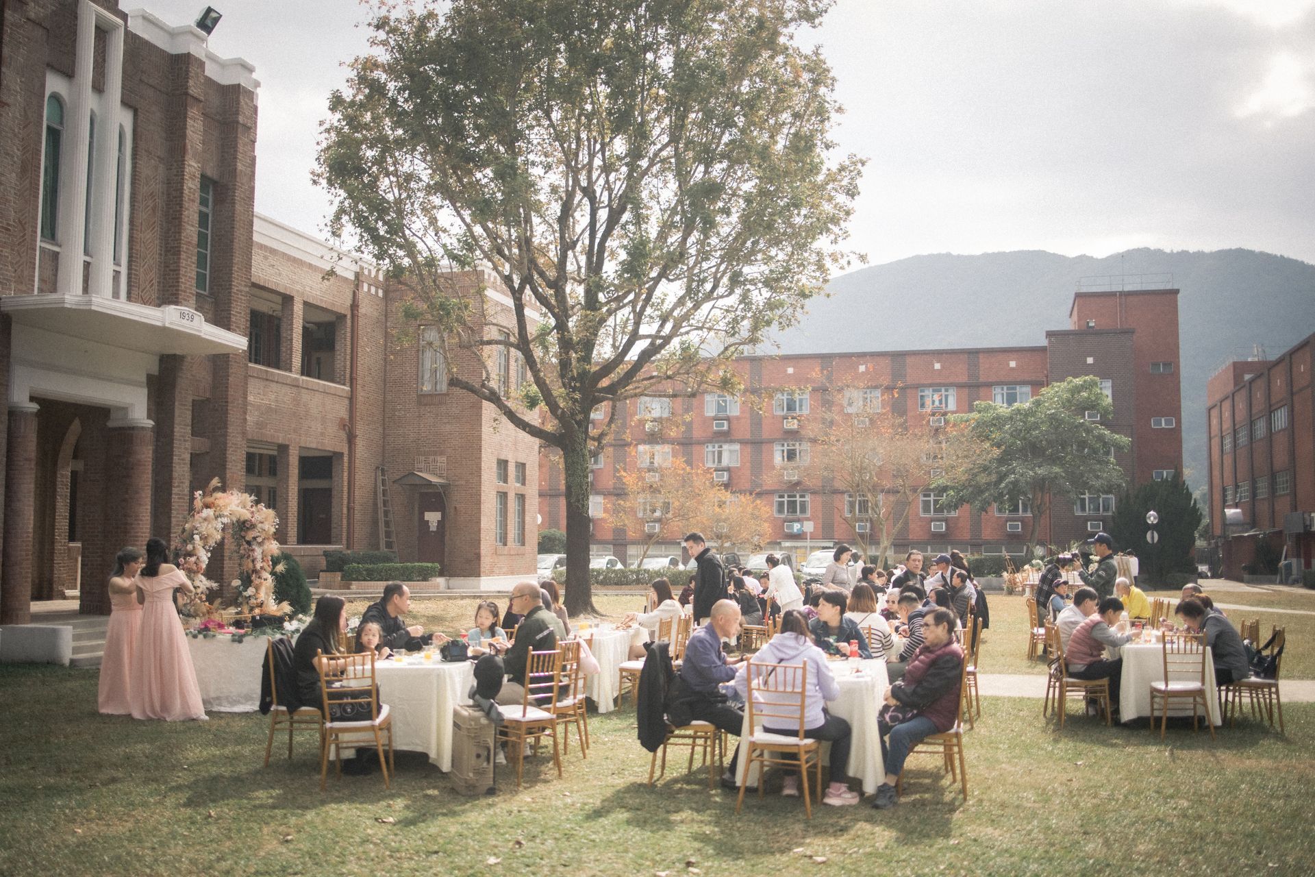 Outdoor wedding reception with guests seated at tables on a lawn near brick buildings and a mountain.
