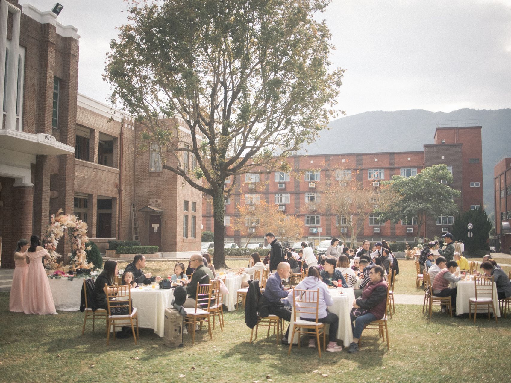 Outdoor event: Tables with guests on a lawn in front of brick buildings, a large tree, and a mountain in the distance.
