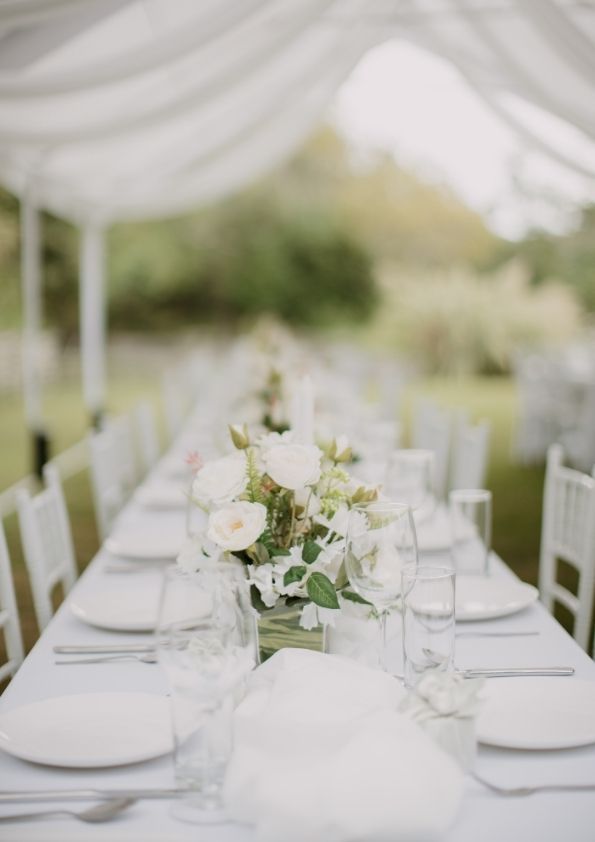 Long white table set for a formal outdoor event, with floral centerpieces and white chairs.