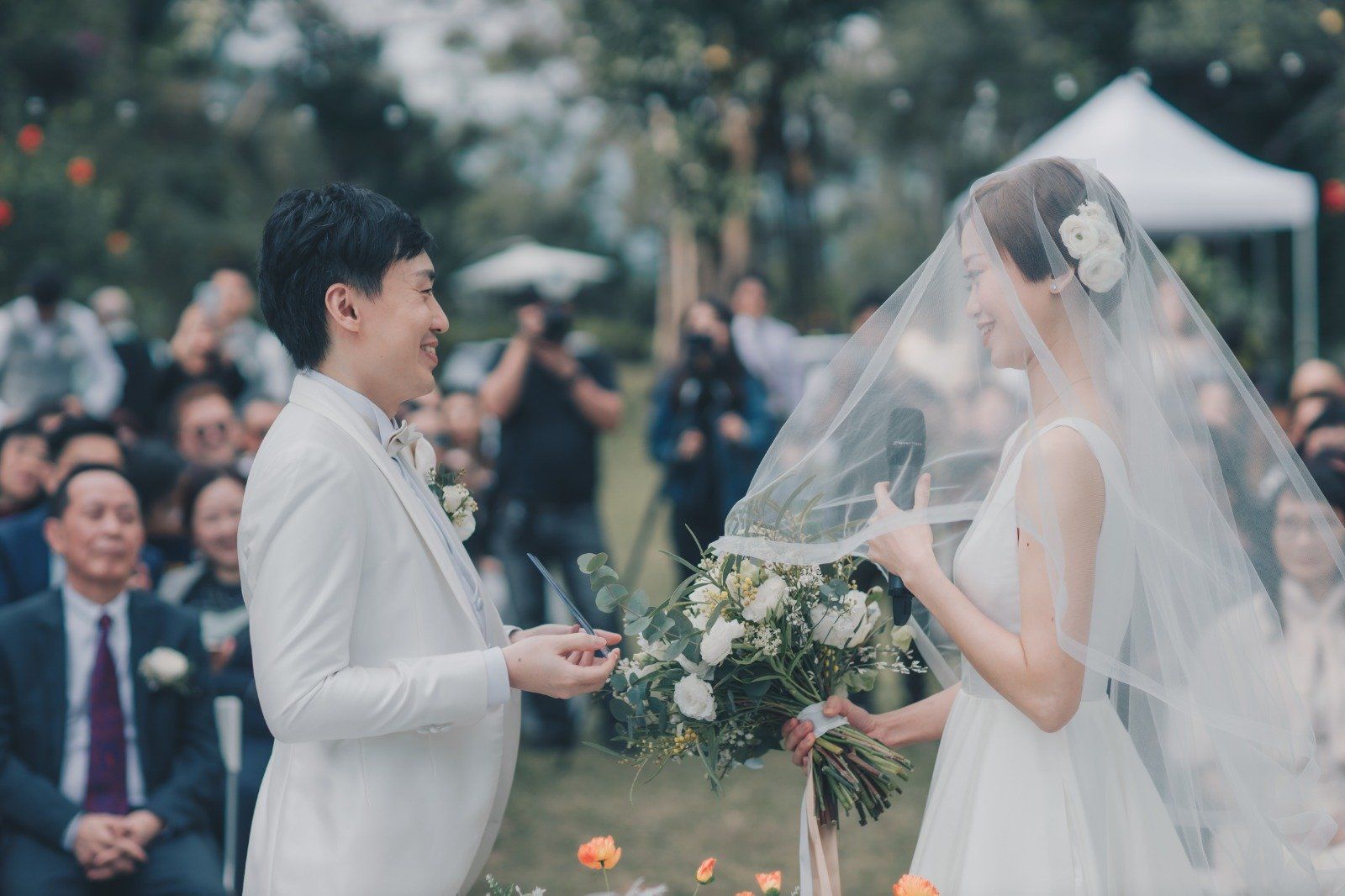 Couple exchanging vows at an outdoor wedding ceremony; bride in veil, groom in white suit, holding flowers.