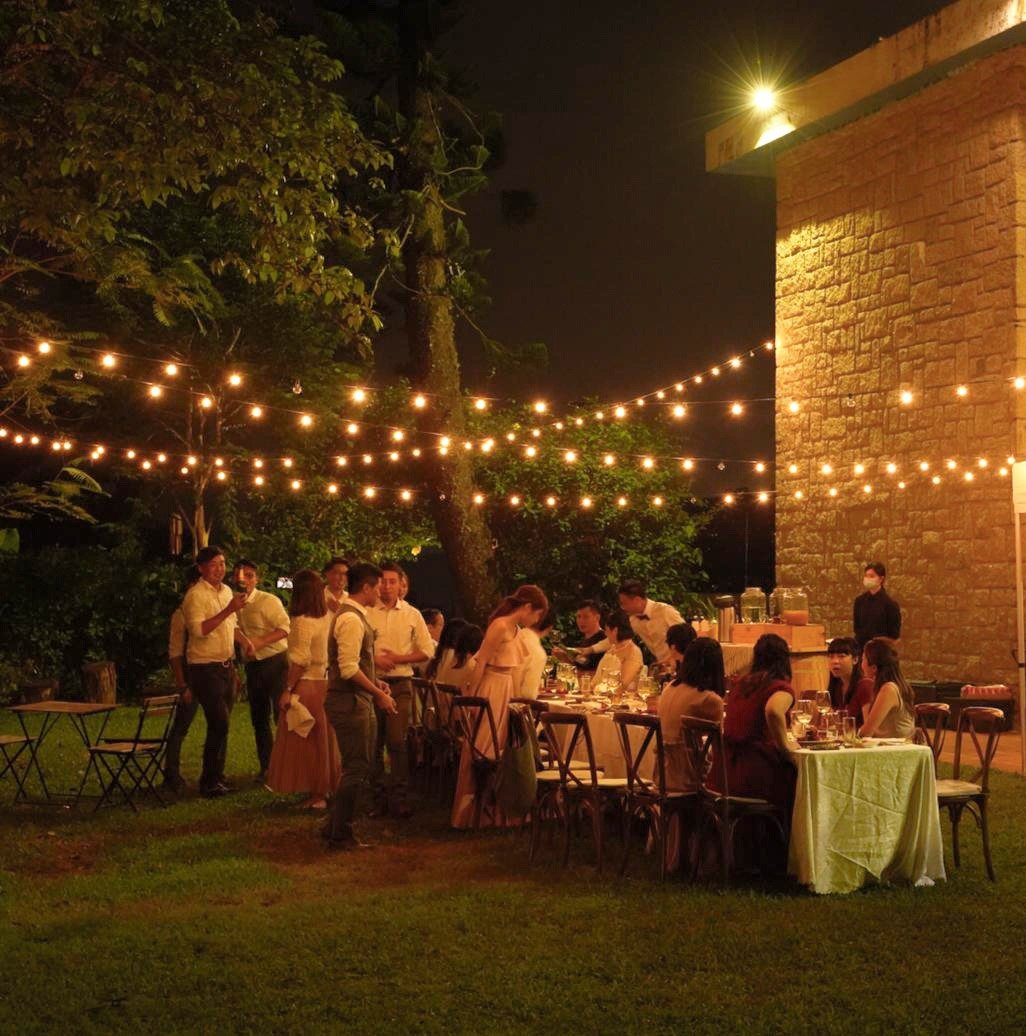 Outdoor dinner party under string lights at night. Tables set with people dining; nearby trees and building.