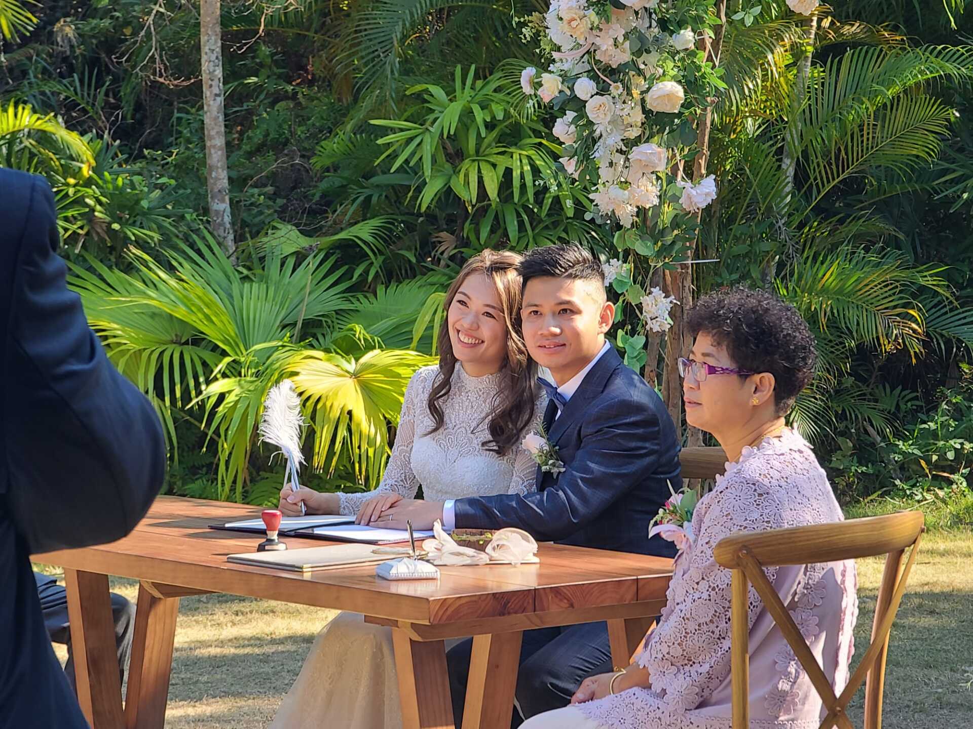 Wedding couple signing documents at outdoor table, guest watches, floral arch.