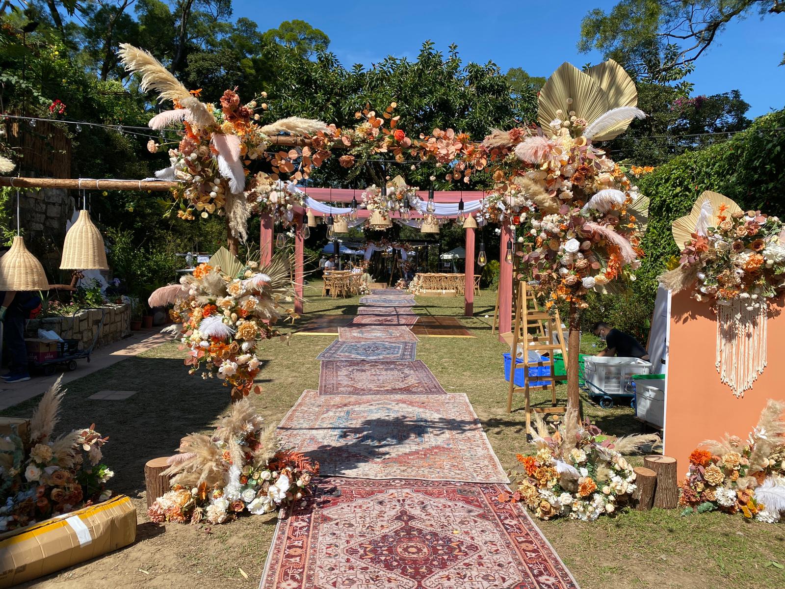 Wedding ceremony setup with floral archway, rug aisle, and decorative arrangements in a garden setting.
