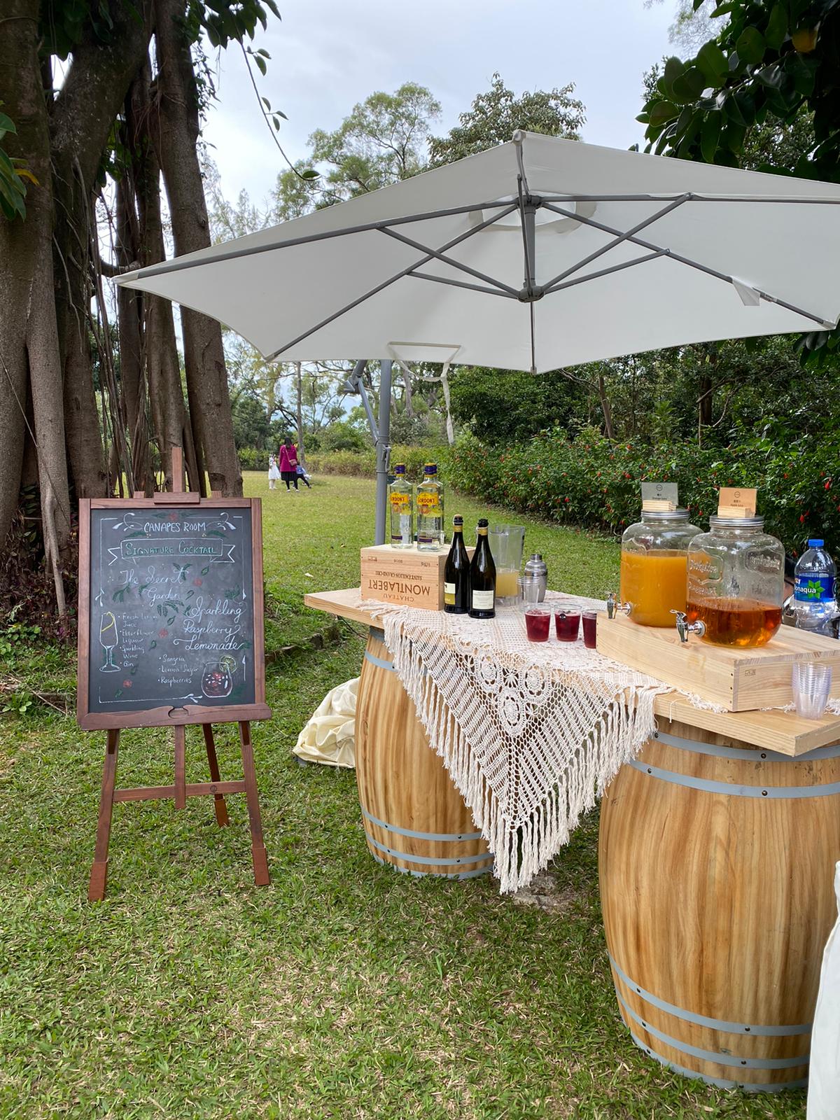 Outdoor bar with wooden barrels, drinks, and a chalkboard sign under a white umbrella.