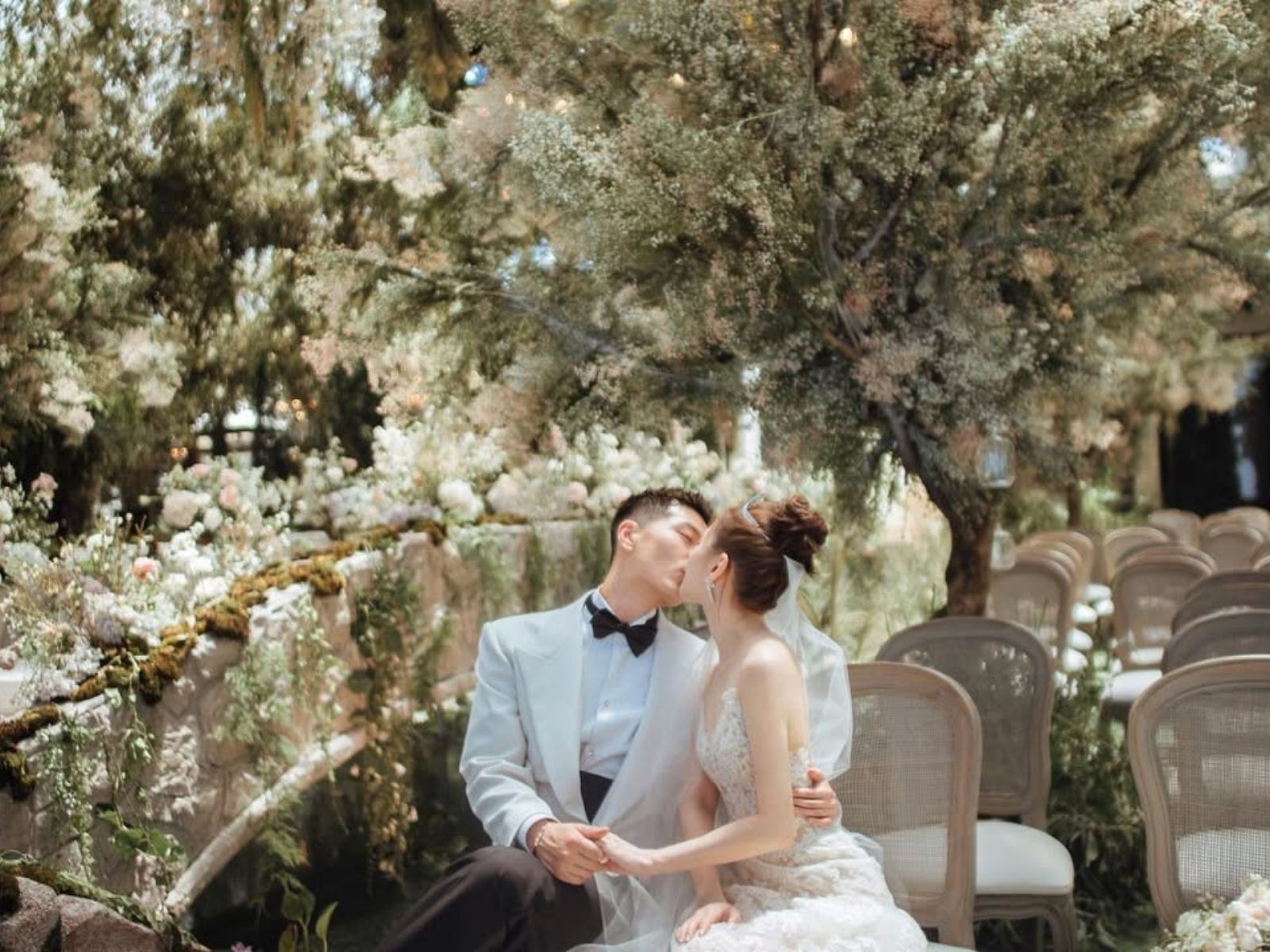 Couple kissing, outdoors wedding ceremony, floral decorations, light colors, seated, romantic.