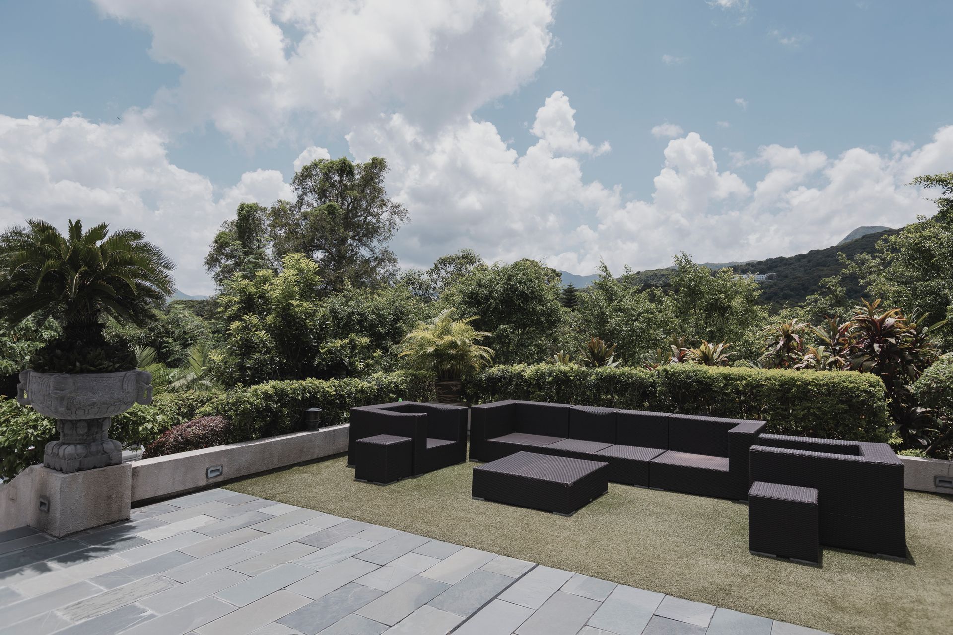 Outdoor patio with black furniture on grass, surrounded by greenery and trees, under a cloudy sky.