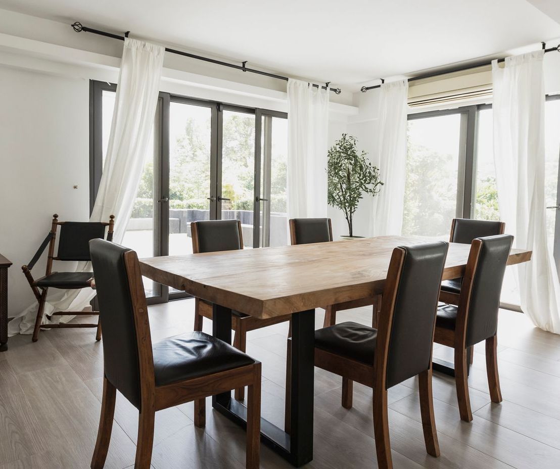 Dining room with wooden table and chairs, large windows with white curtains.