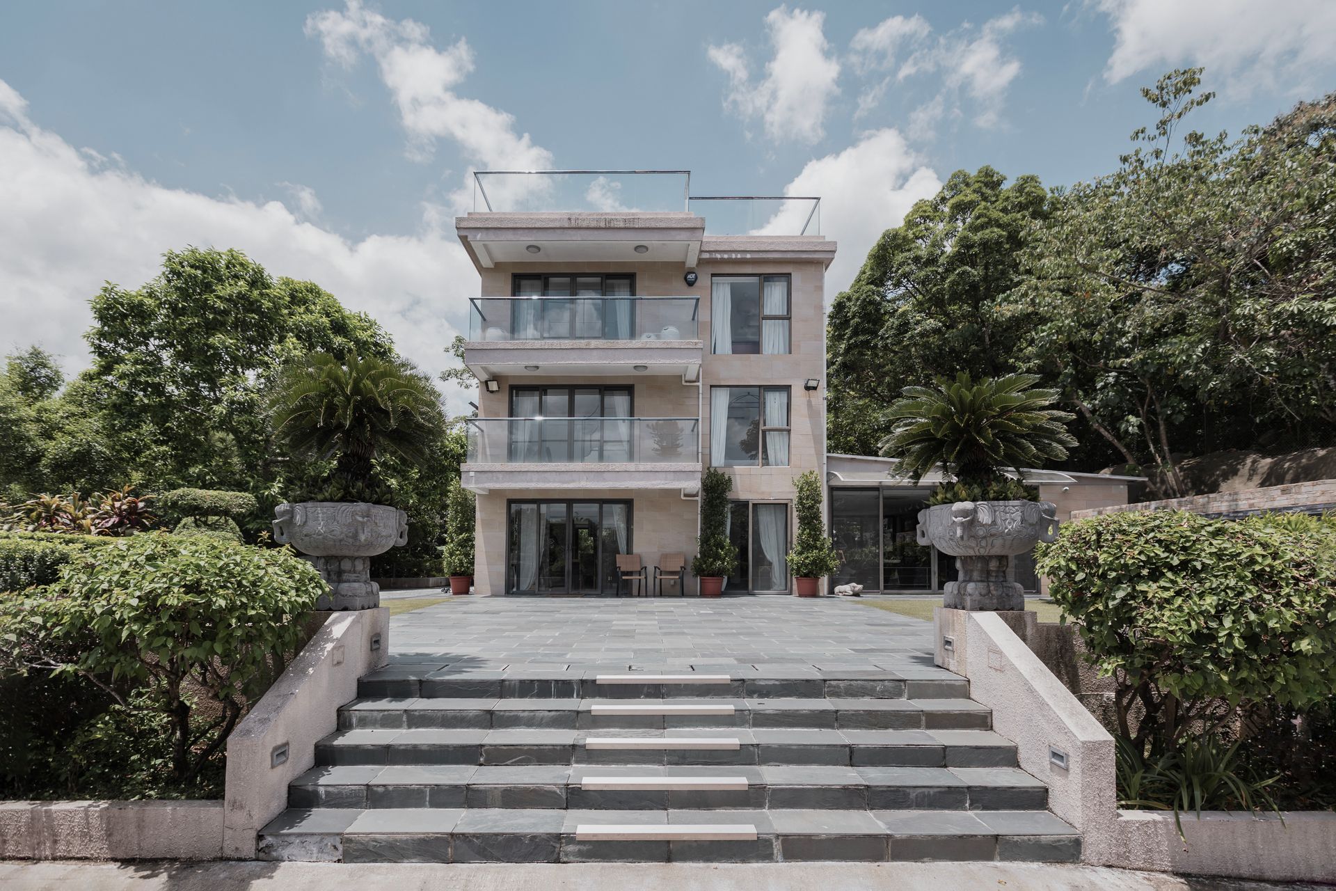Three-story house with glass balconies, accessed by stone steps, nestled among green trees under a cloudy sky.