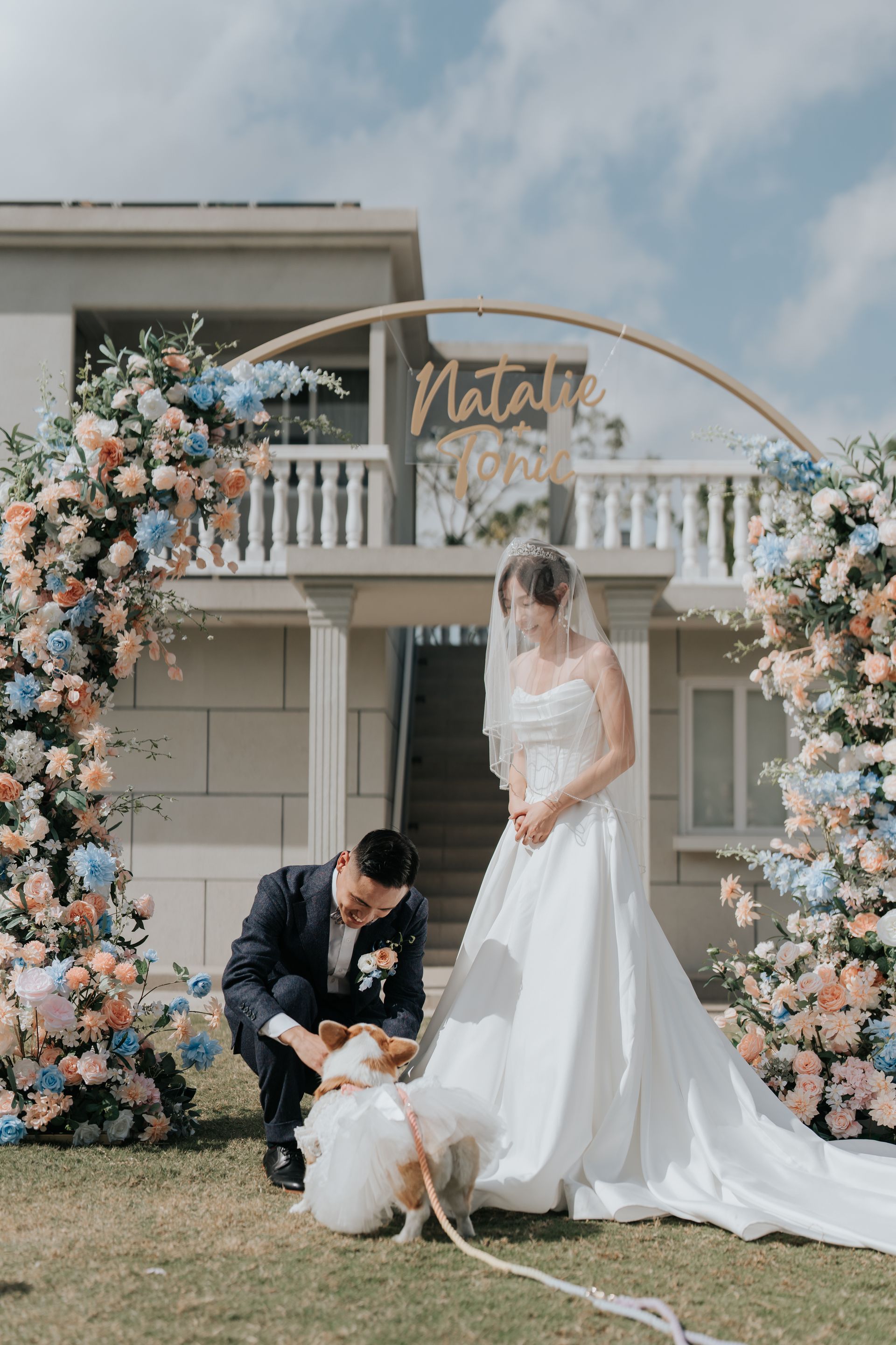Wedding: Bride and groom with dog in front of floral arch, house in background.