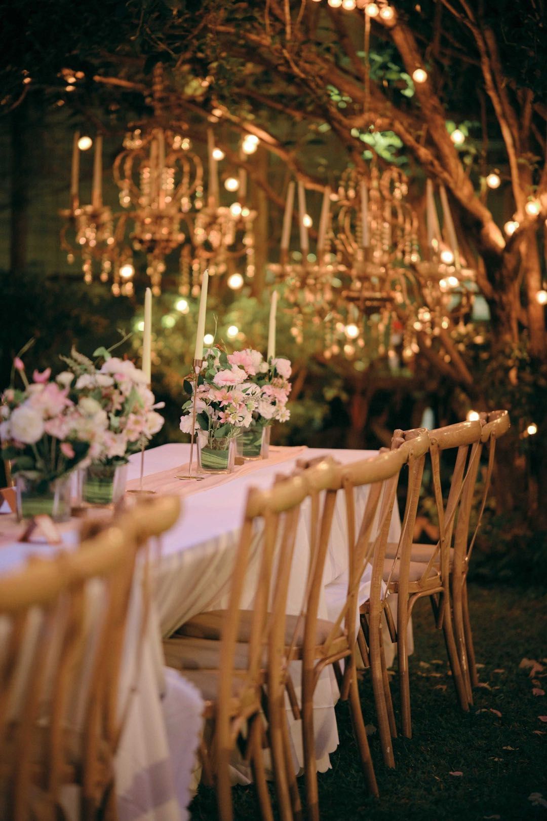 Outdoor wedding reception table, decorated with flowers and chandeliers, set under a tree with soft lighting.