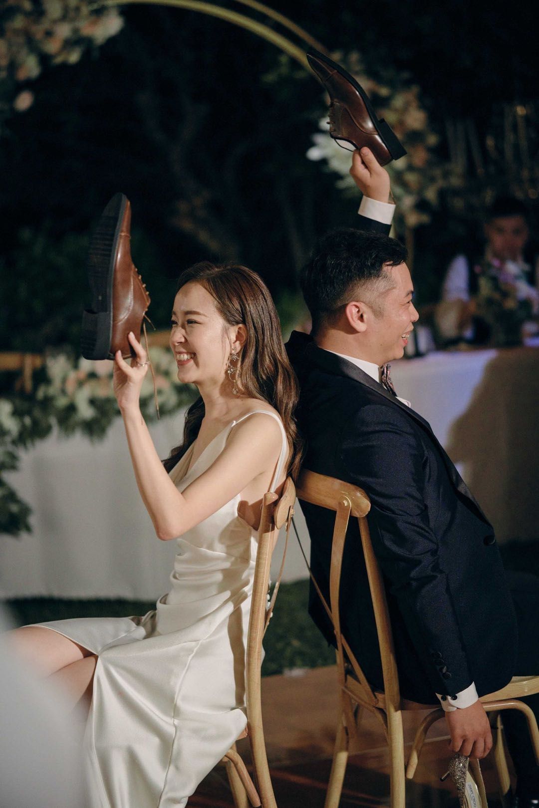 Newlyweds play shoe game: smiling, holding shoes above heads, seated back-to-back at an outdoor wedding.