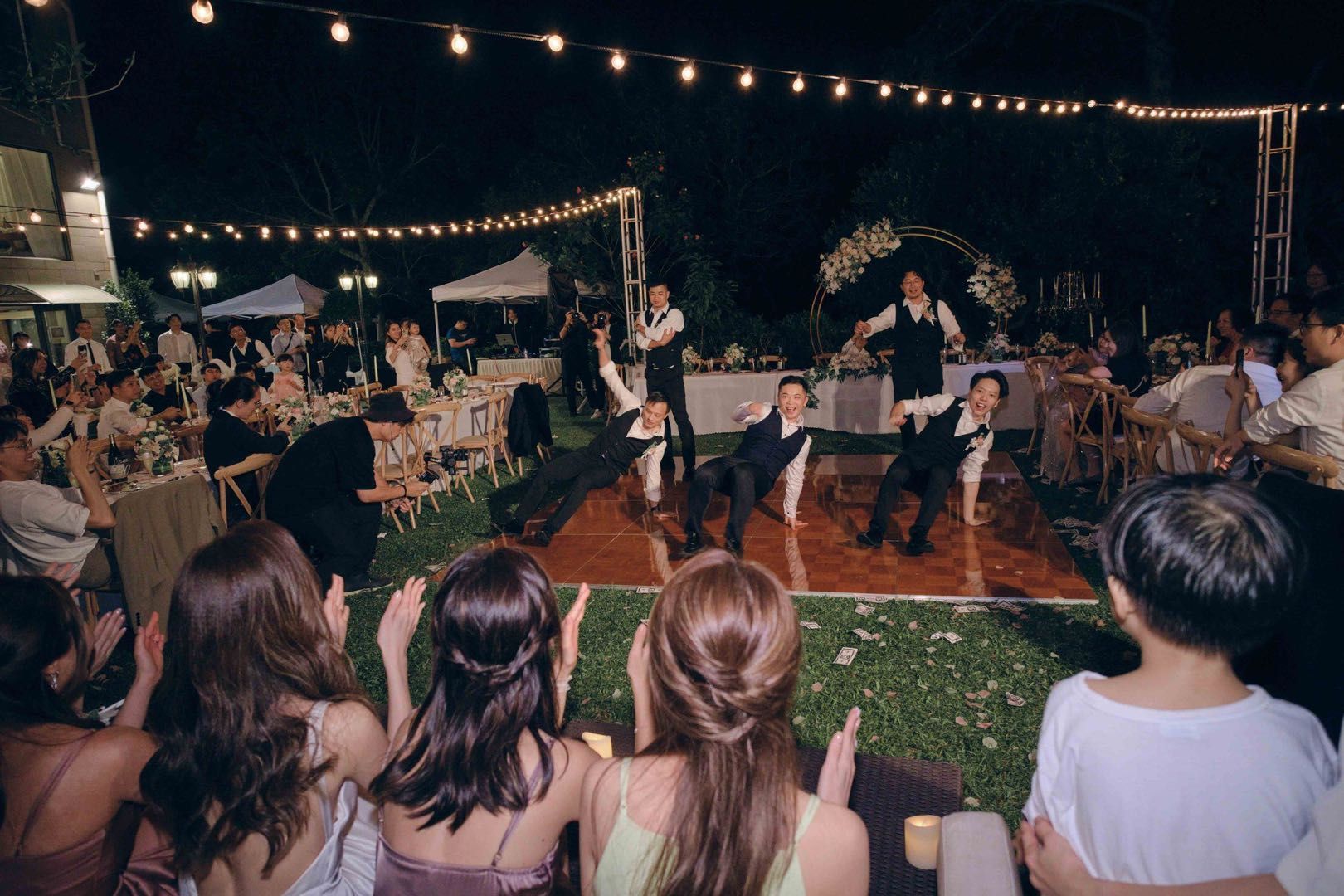 Wedding reception: Guests watch men in vests perform a dance on a lit dance floor under string lights.