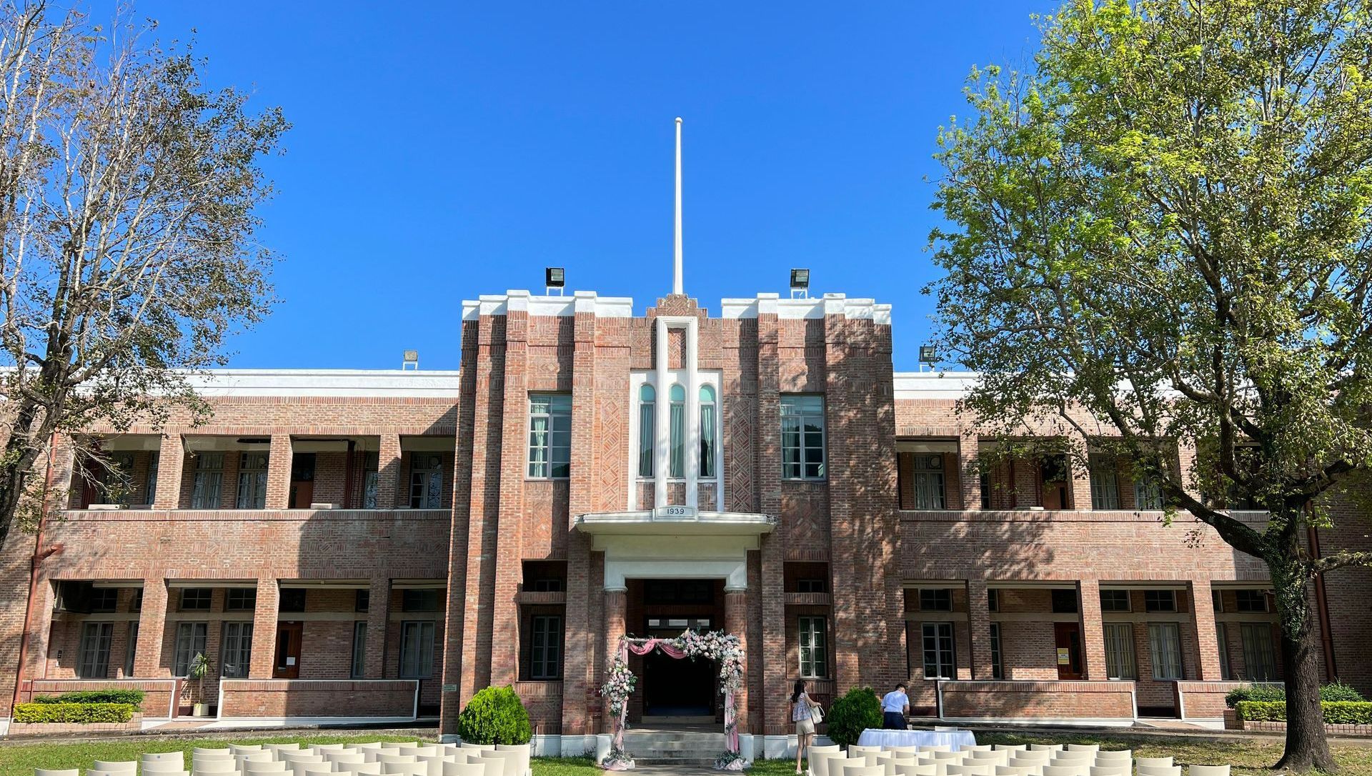 Brick building with a white central entrance, under a clear blue sky. Empty white chairs on grass.