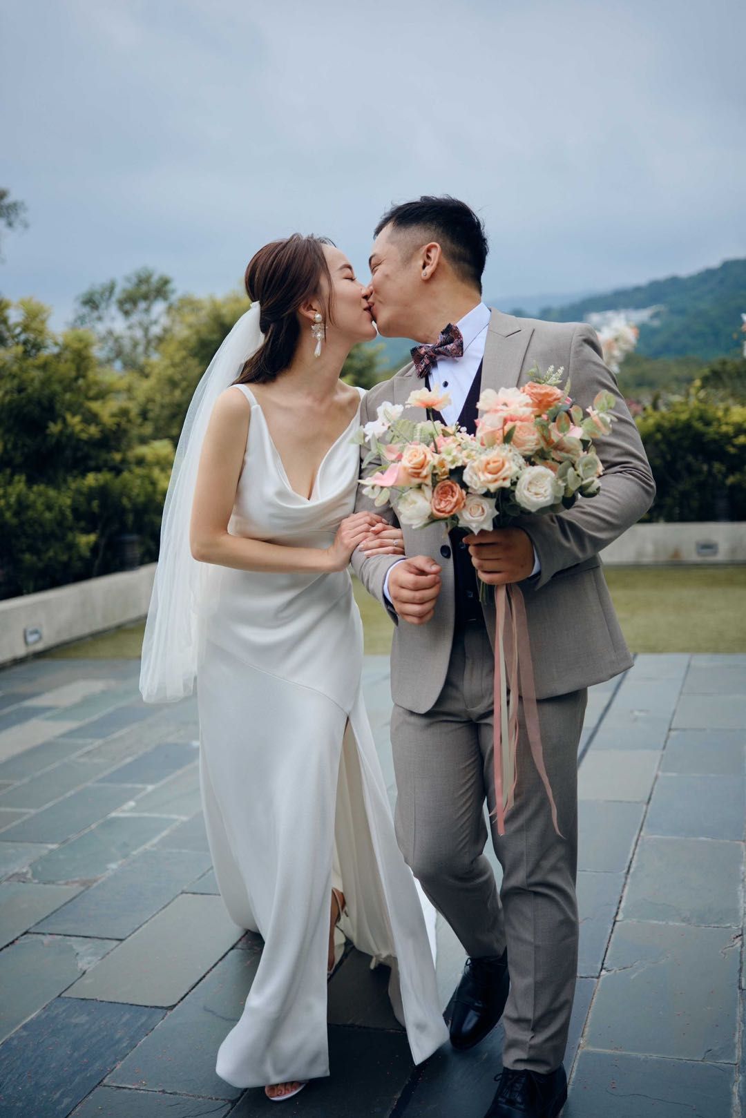 Newlyweds kiss, bride in white dress, groom in grey suit, holding bouquet, outdoor setting.
