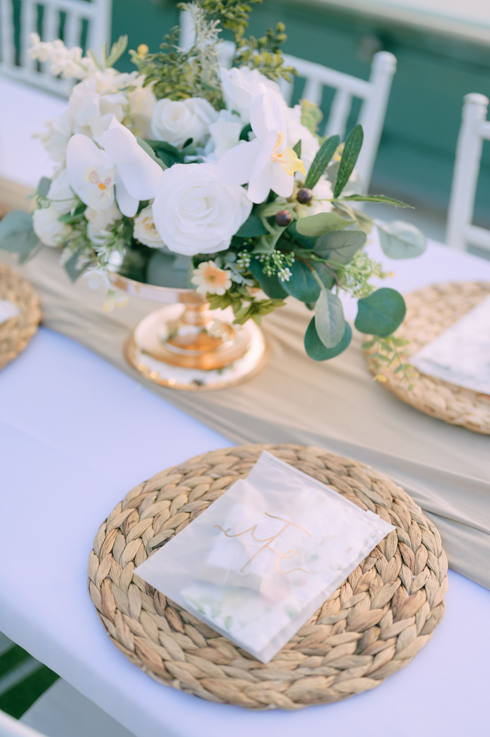 Table setting with white flowers, gold accents, woven placemats, and a beige runner.
