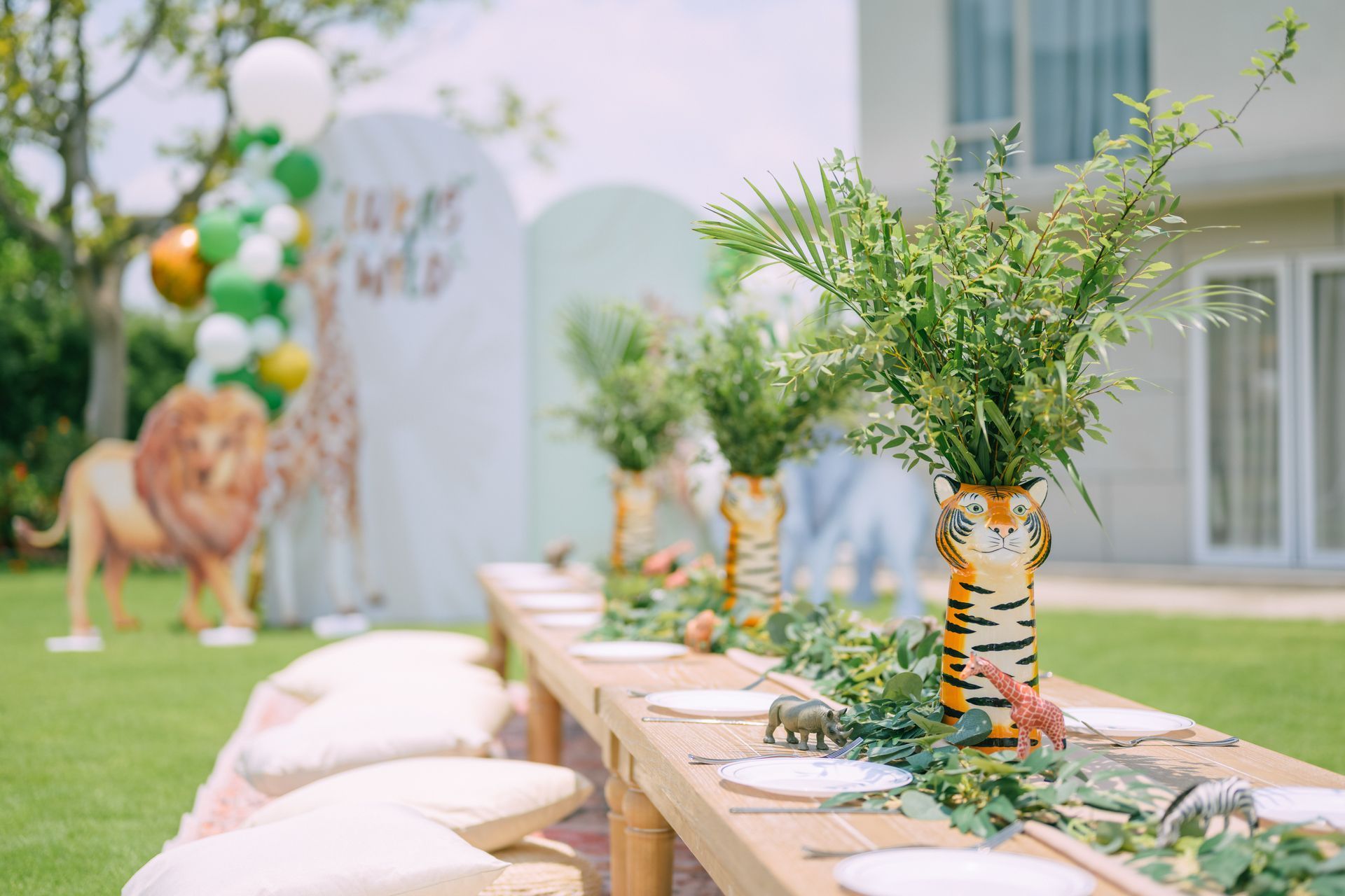 Outdoor decorated aisle with white cushions, greenery, and tiger-striped wrapped vases near a building