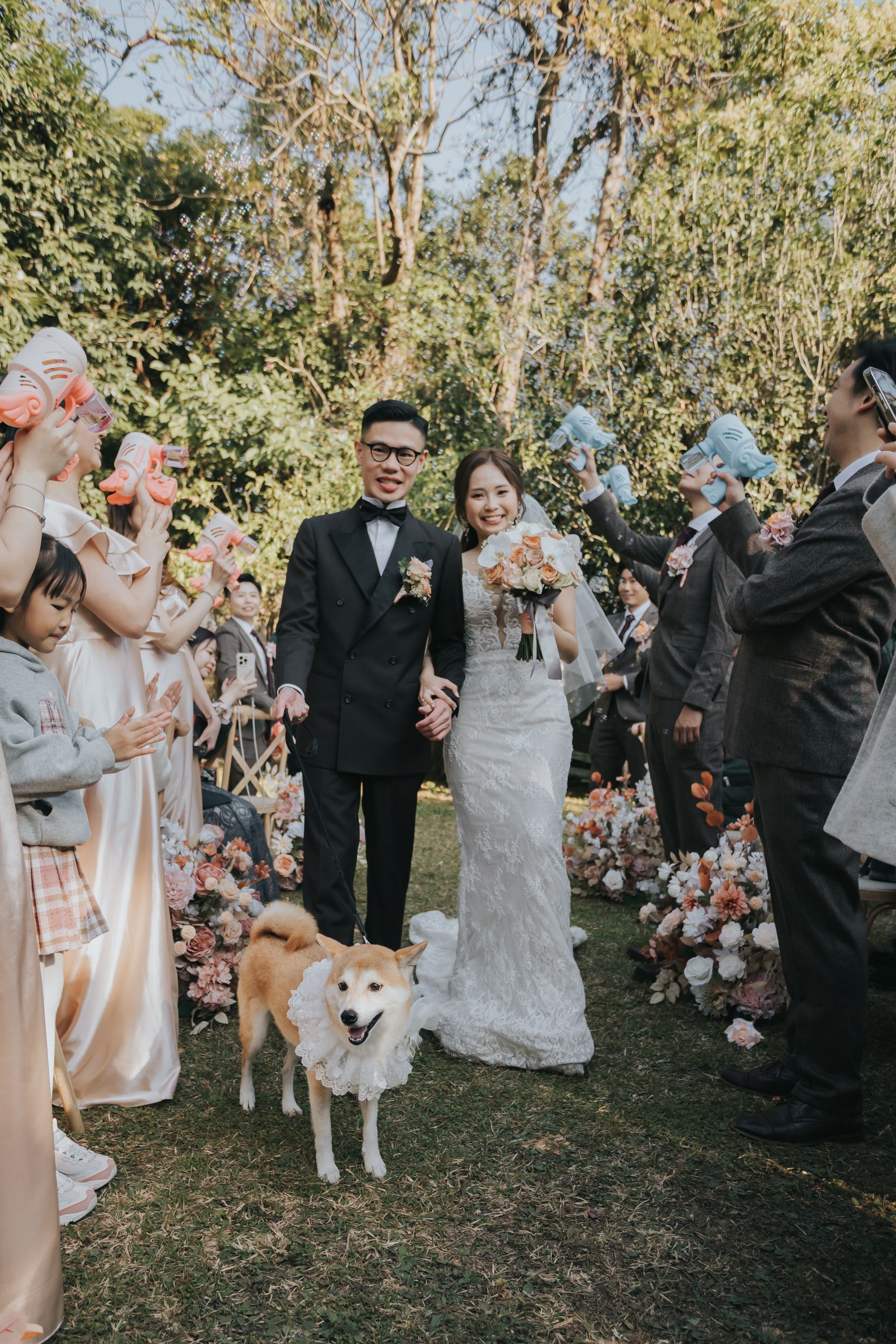 Newlyweds walk through an outdoor garden, holding hands, followed by wedding party and a dog with a ruffled collar.