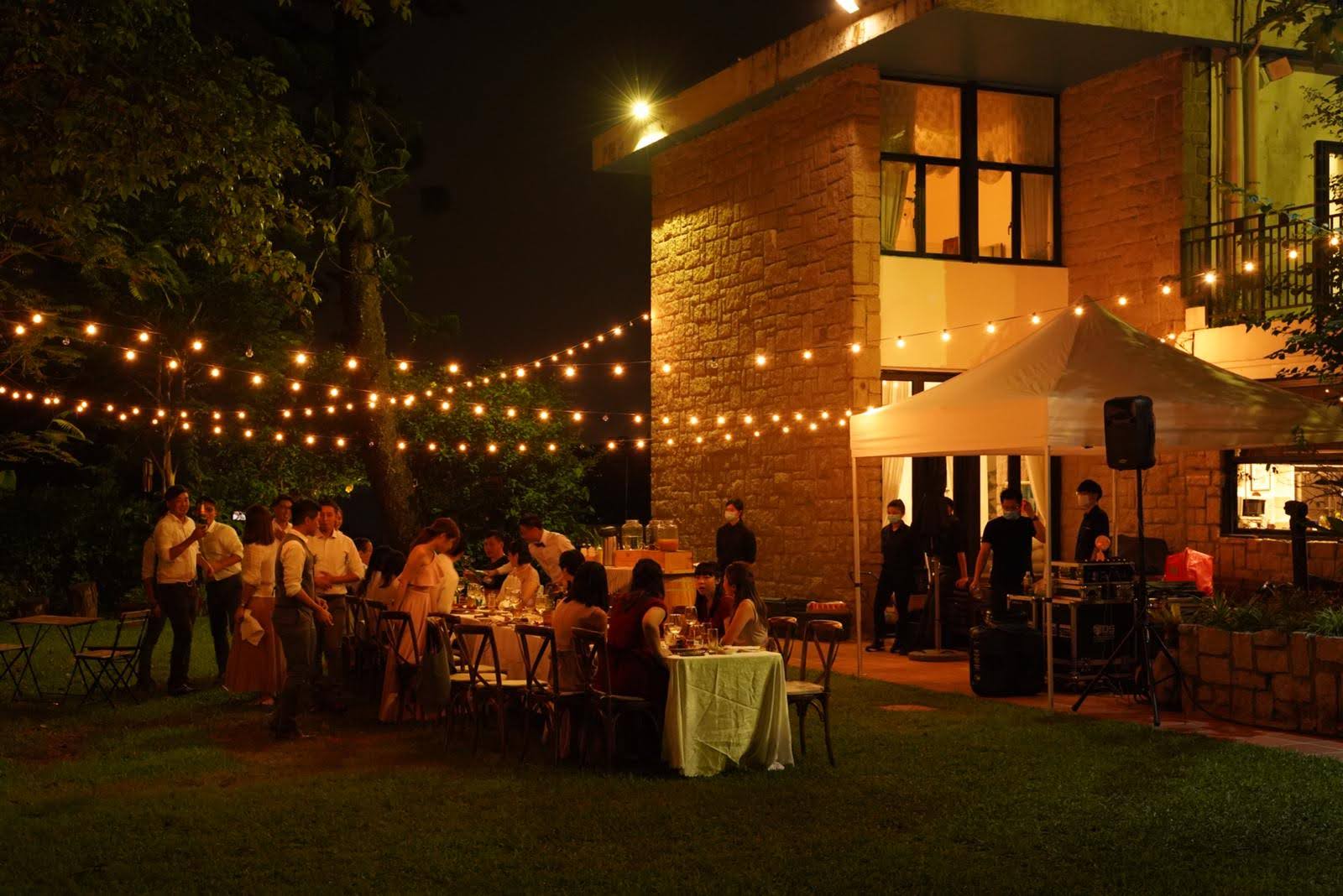 Outdoor evening gathering with string lights, tables, and people near a building.