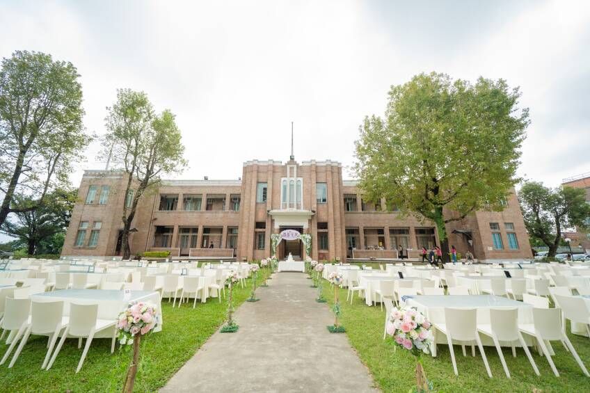 Wedding reception set up on lawn, tables and chairs facing a decorated arch in front of a brick building.