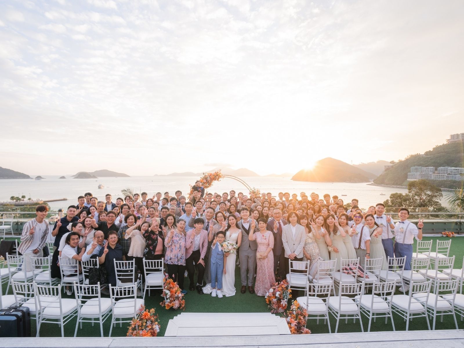 Large group at an outdoor wedding ceremony, overlooking the ocean at sunset.