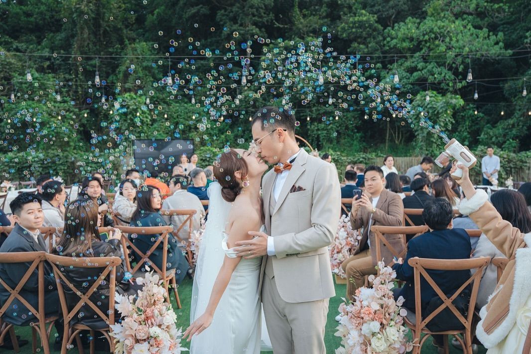 Bride and groom kissing at outdoor wedding ceremony; bubbles and guests surround them.