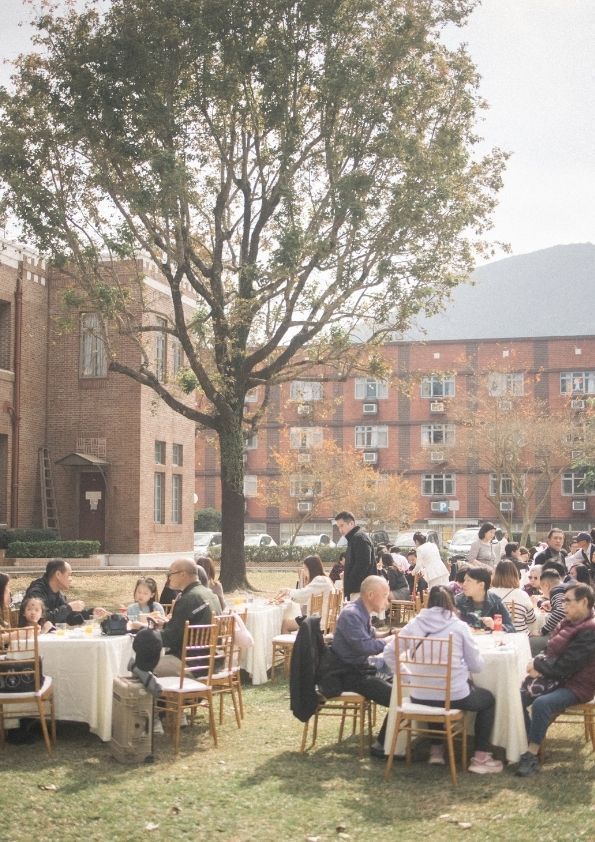 Outdoor event with people seated at tables on grass, near brick buildings and trees.