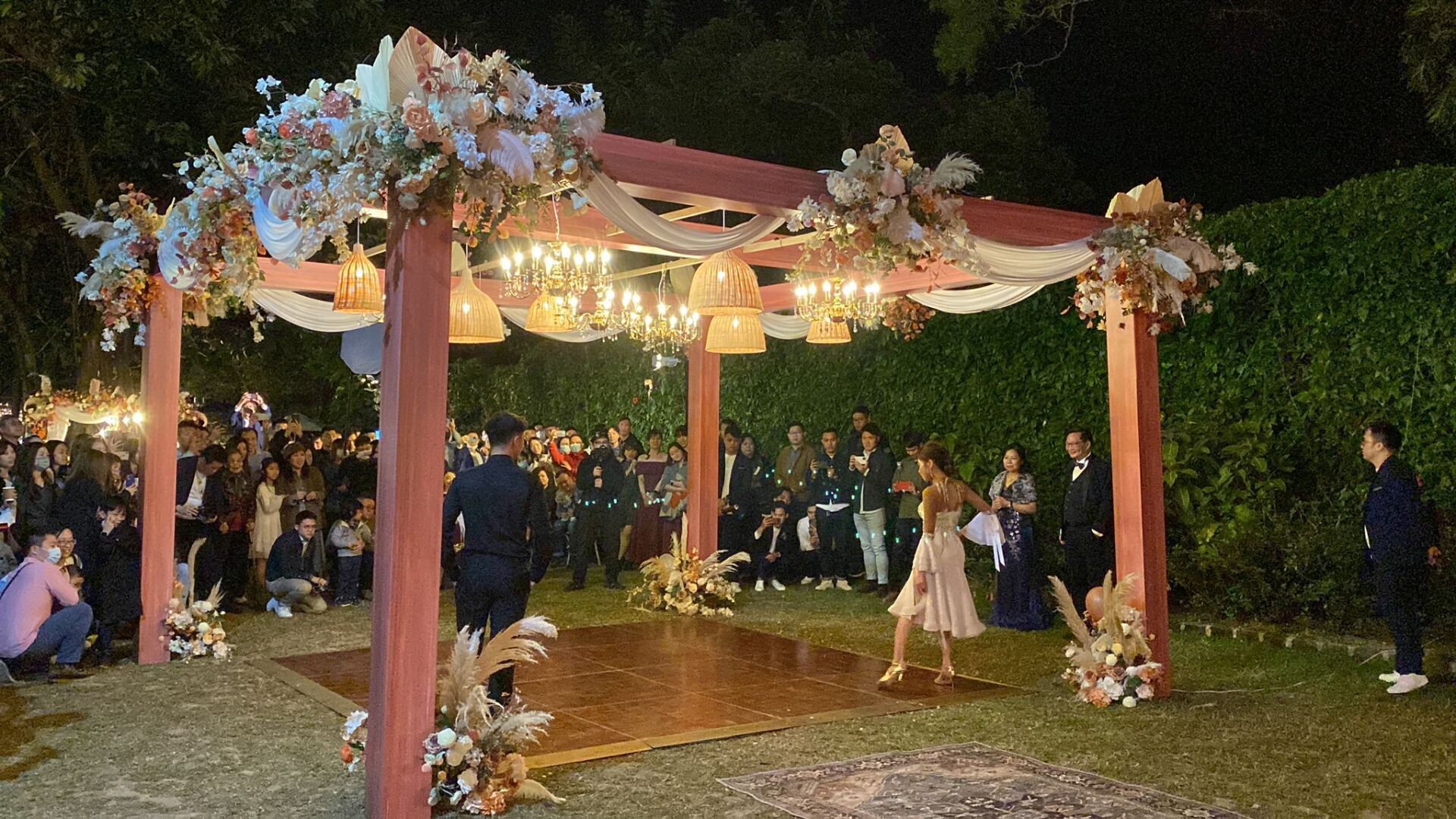 Wedding celebration with a decorated wooden canopy, dance floor, and guests in an outdoor setting at night.