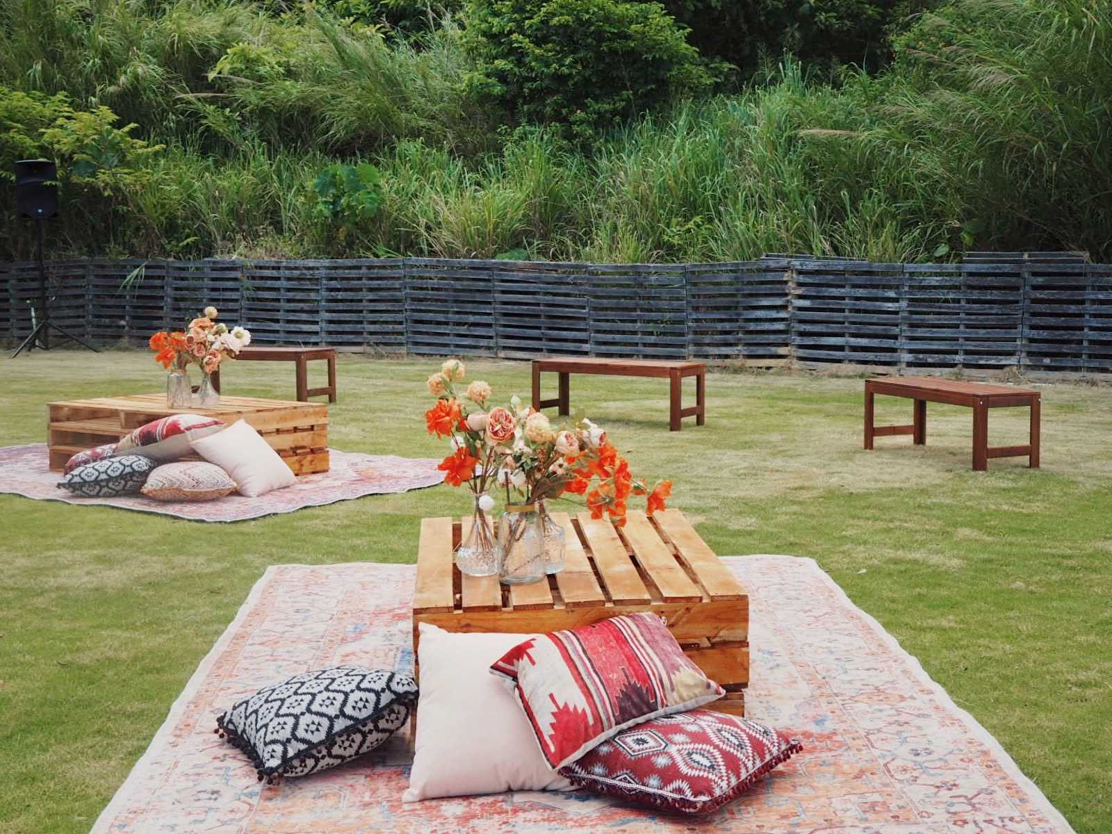 Outdoor seating area with low wooden tables, cushions, and rugs on green grass. Flowers in vases.