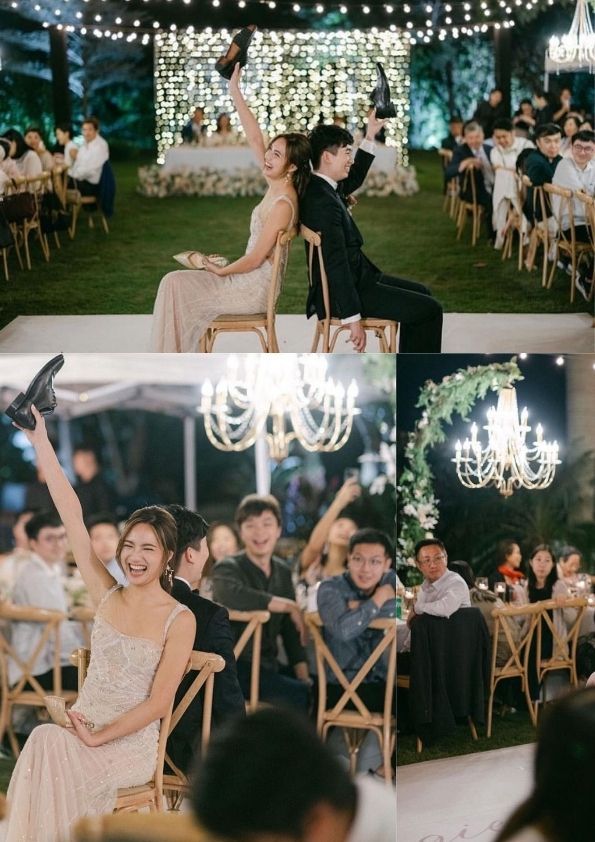 Wedding game: Bride and groom back to back, holding up shoes. Guests watch, laughing. Outdoor reception.