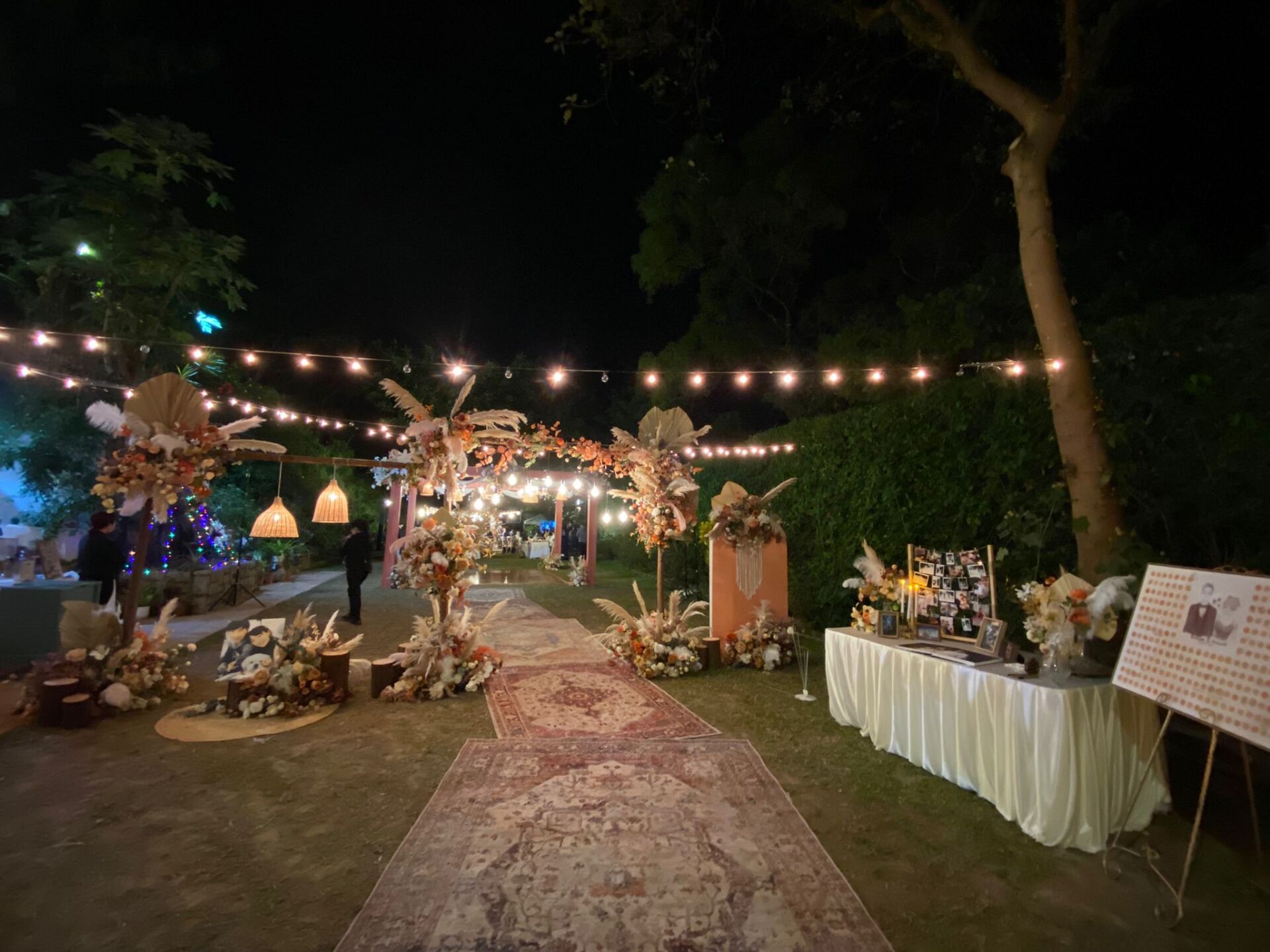 Outdoor evening event with string lights, floral arrangements, and a rug leading to a buffet table.