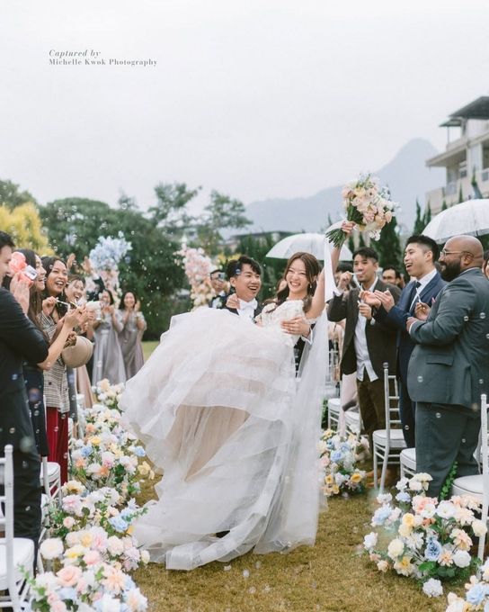 Groom lifting bride at outdoor wedding reception. String lights and guests in background, mountain view.