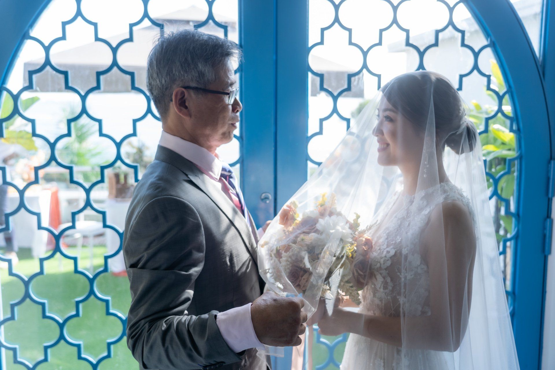 Father adjusting bride's veil before wedding ceremony, standing near blue, ornate window.