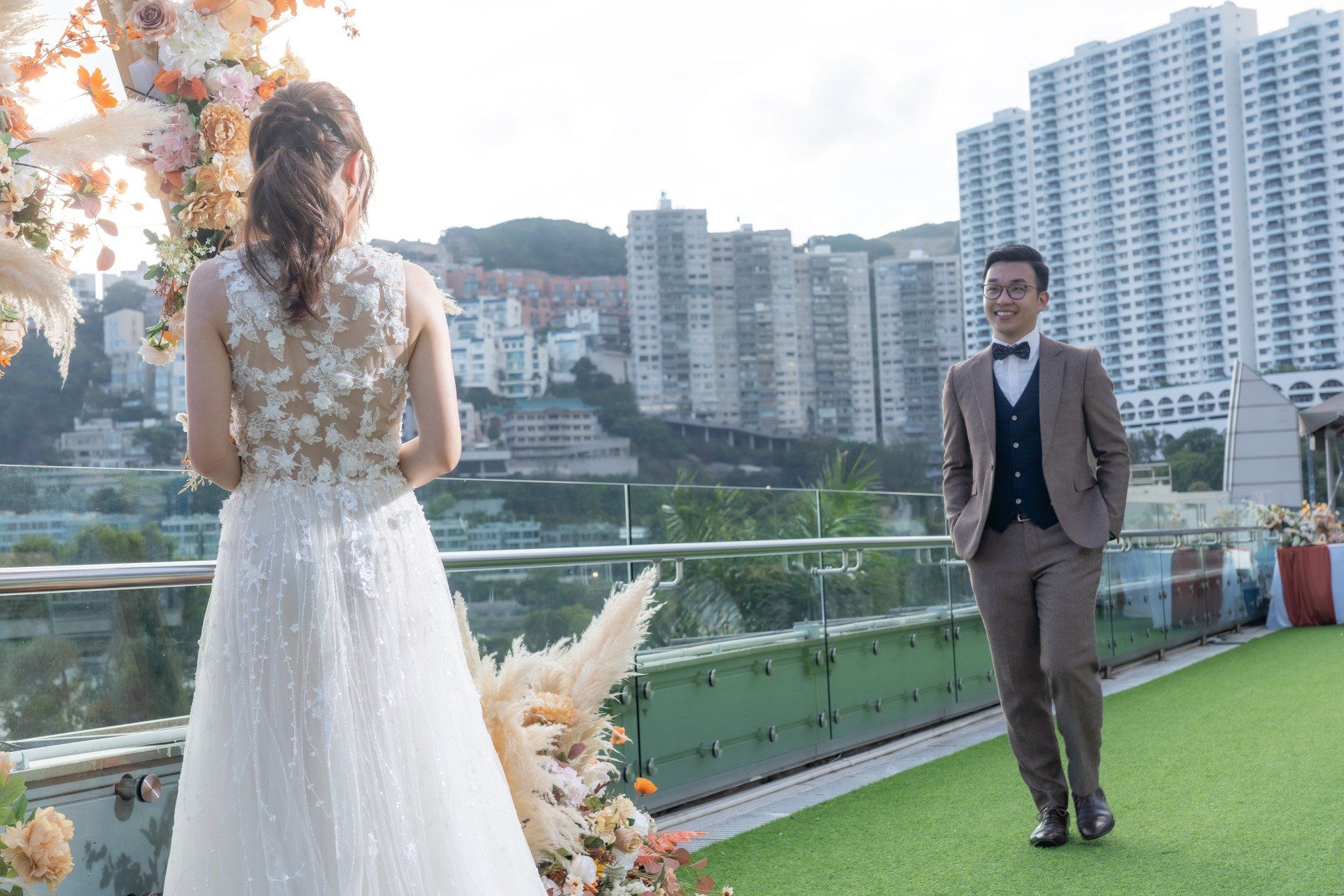 Bride facing away, groom smiling. Wedding scene on rooftop with cityscape.