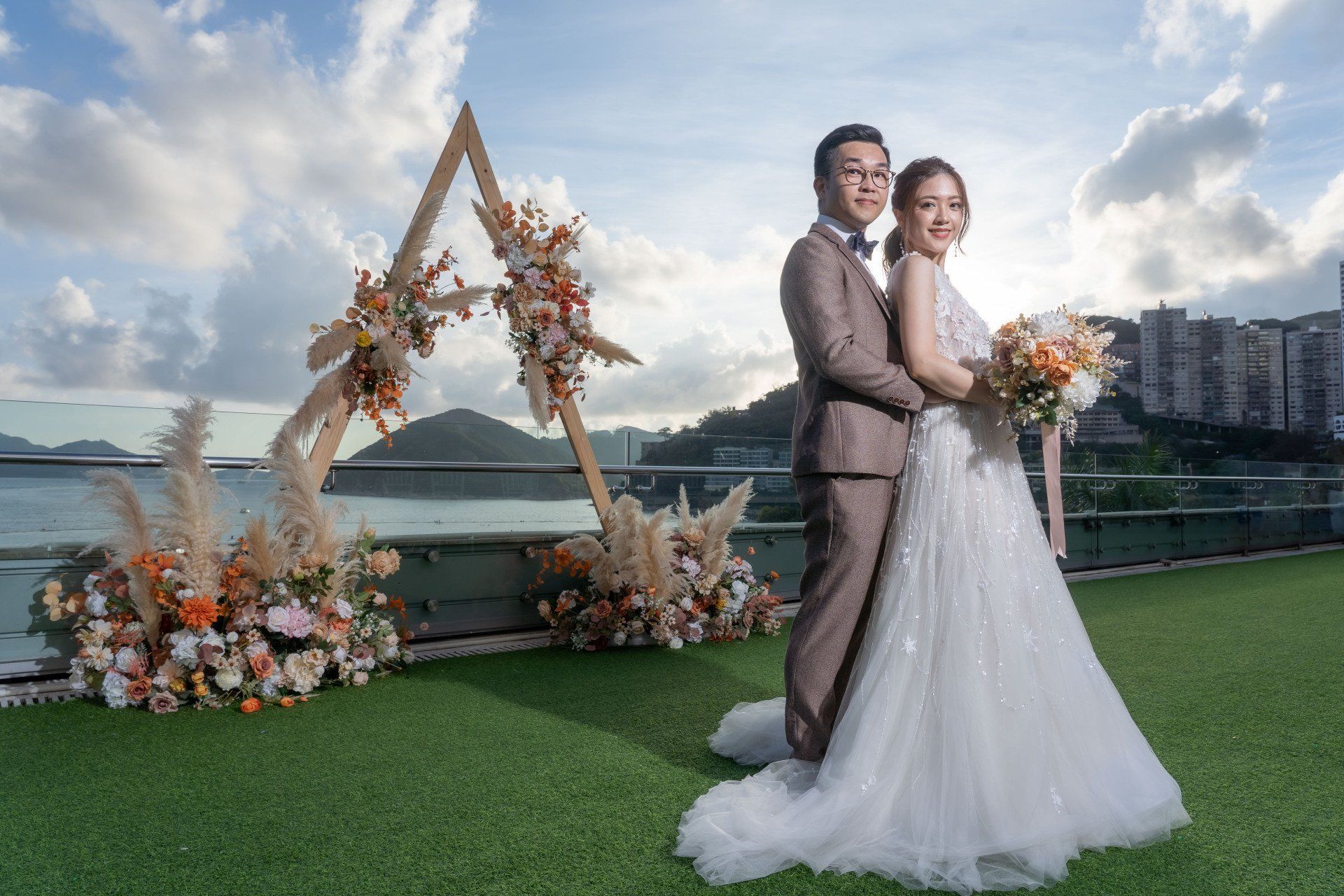Newlyweds pose outdoors, near a wooden arch decorated with flowers. Man in brown suit, woman in white dress.