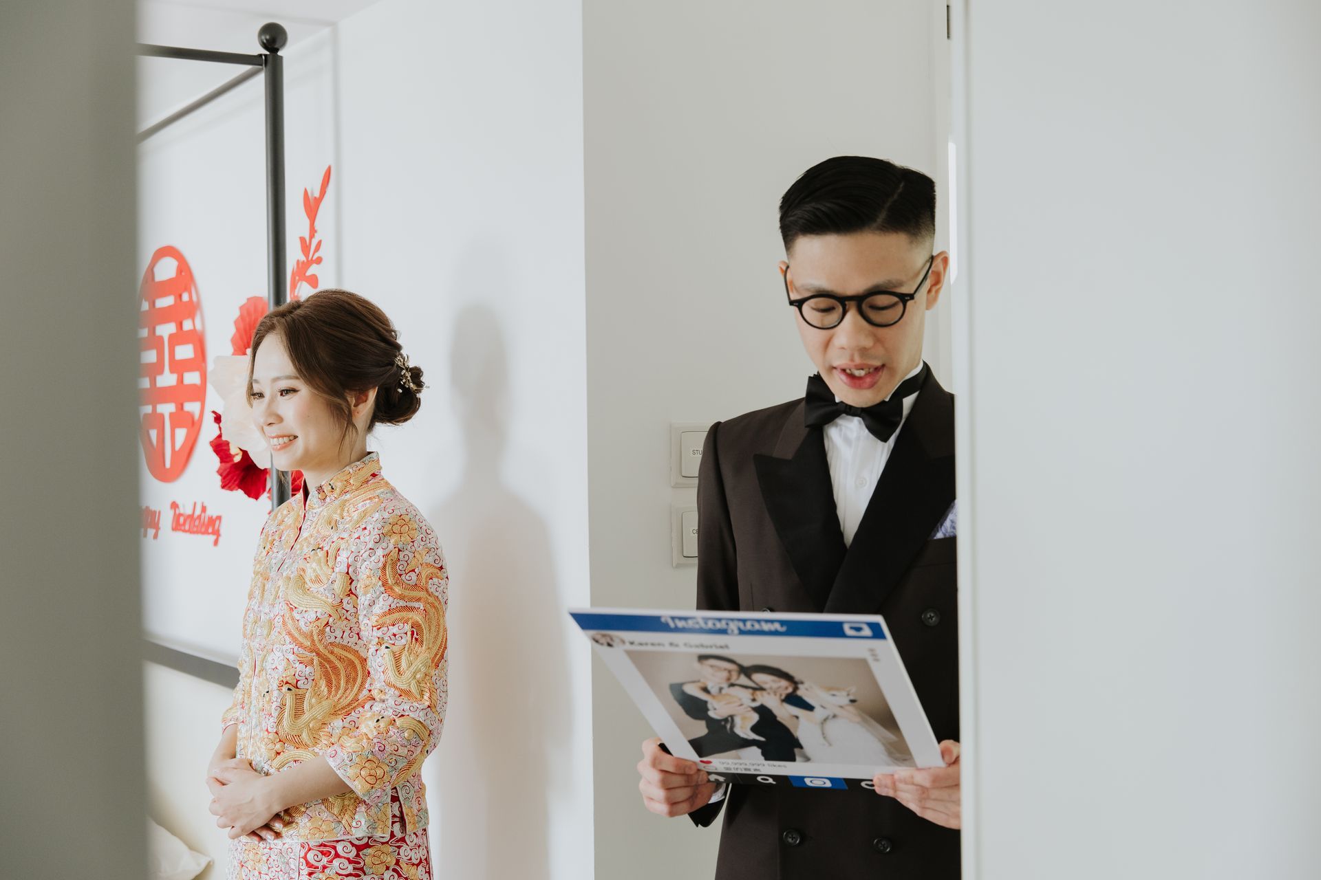 Groom in tuxedo holds framed photo, smiling at bride in traditional dress in doorway.