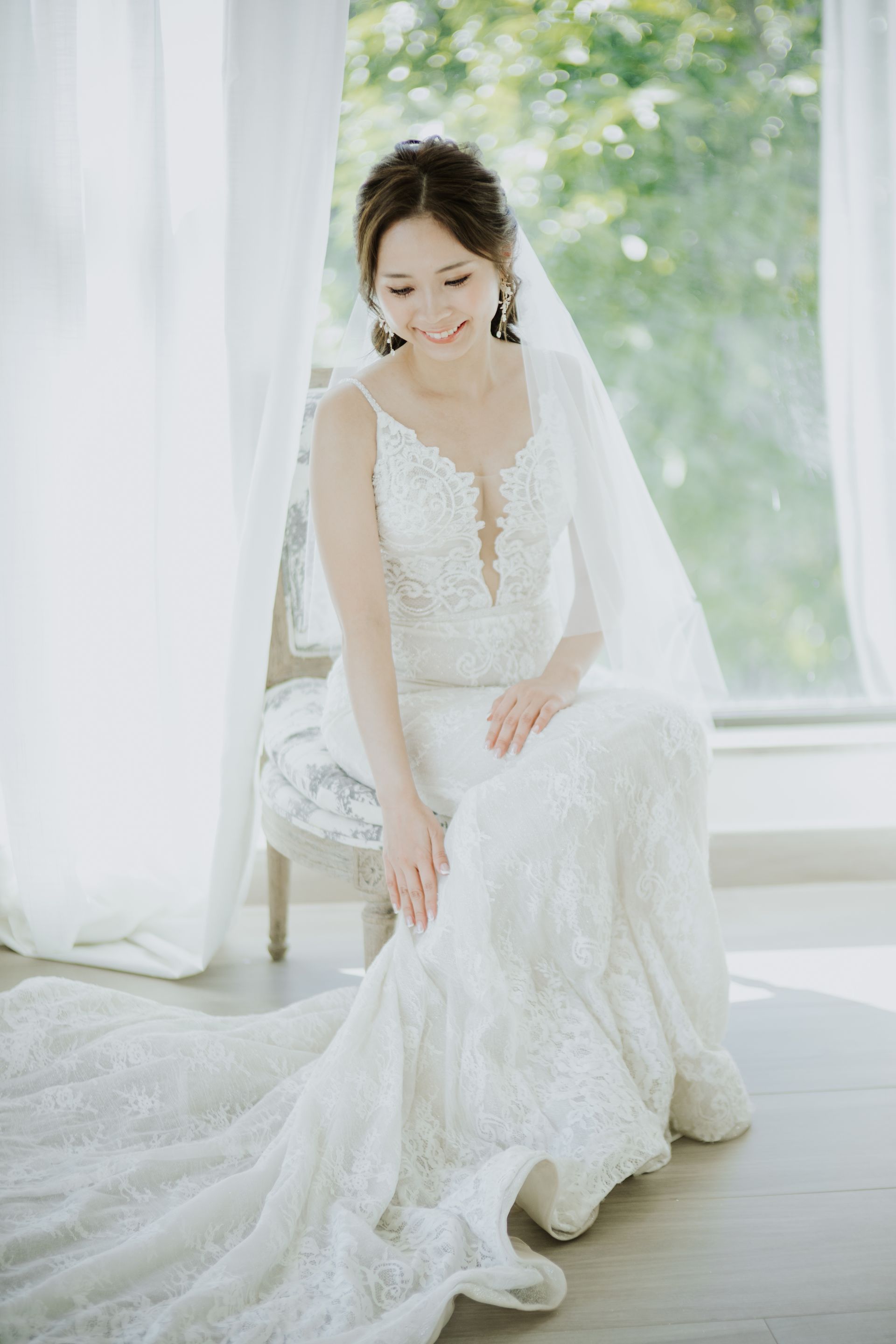 Bride in a white lace wedding dress, veil, seated, smiling near a window.