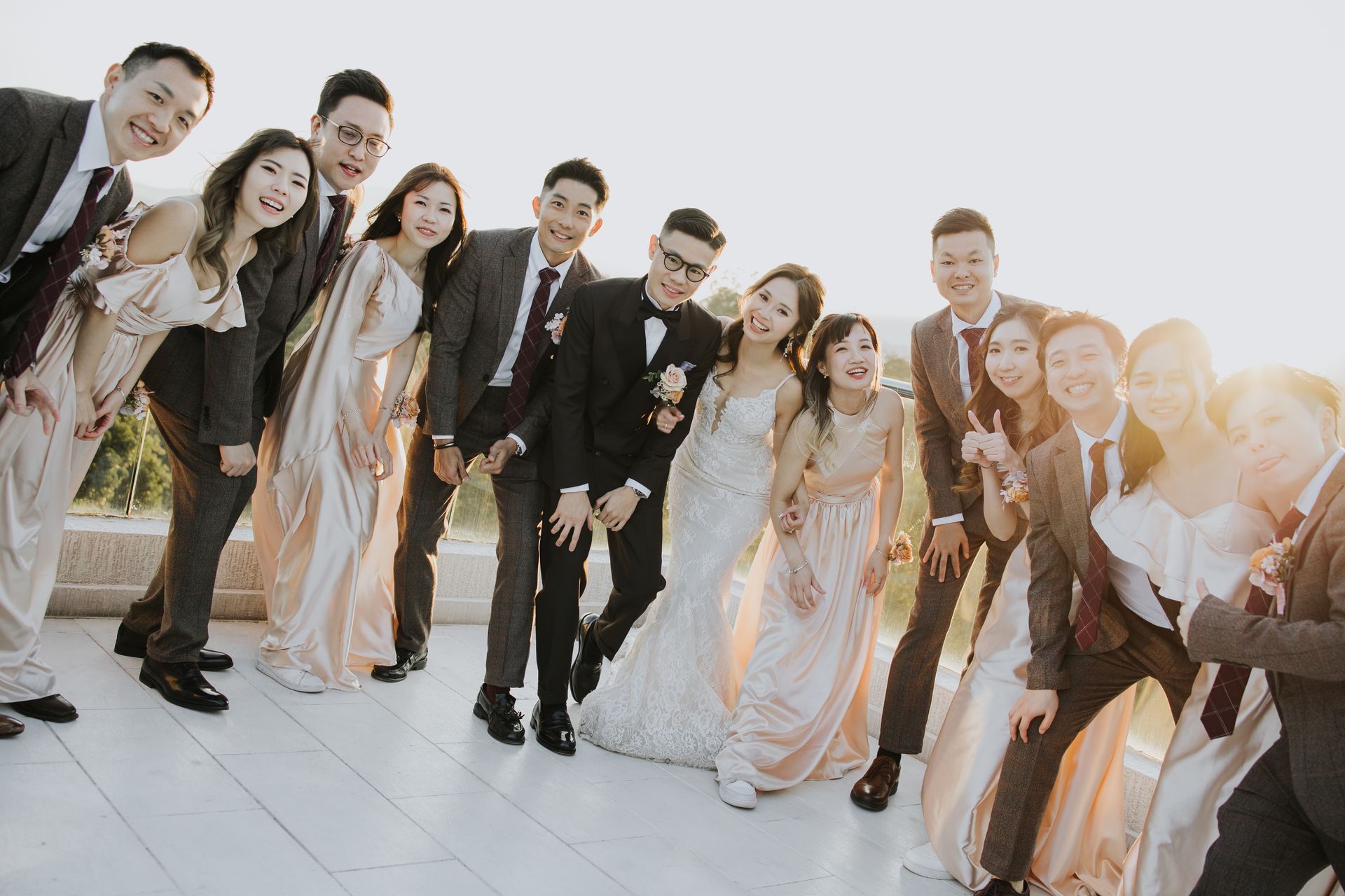 Wedding party poses on a rooftop: bride and groom, bridesmaids in peach, groomsmen in suits, smiling in sunlight.